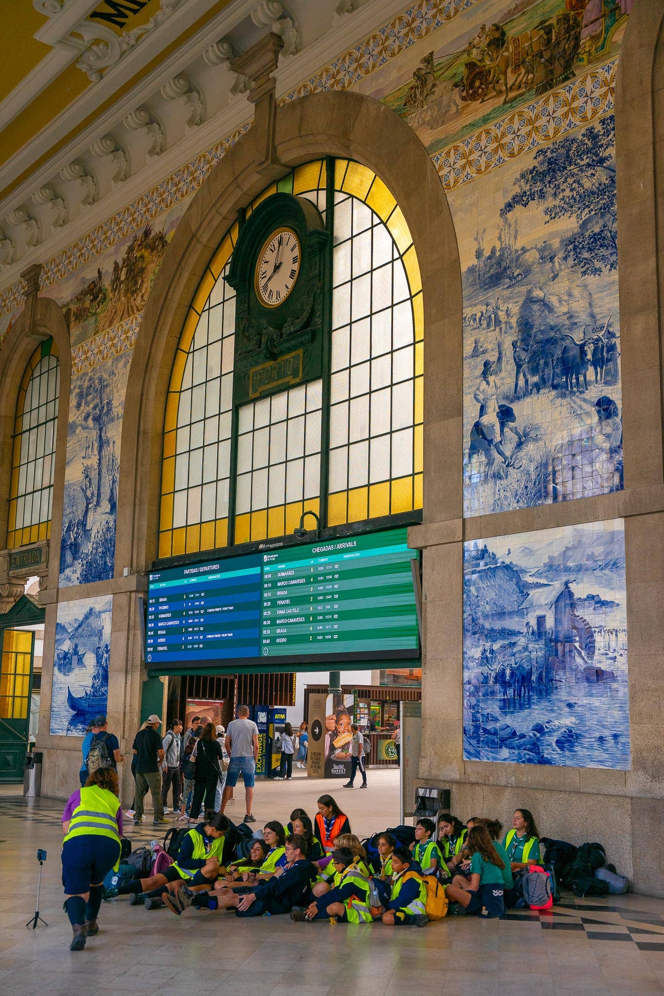 The concourse of Sao Bento station in Porto, its walls covered in blue-and-white azulejo panels beneath an arched window and a departures board.