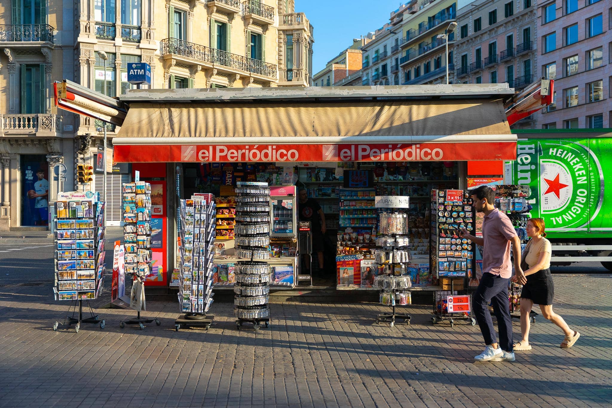 A newspaper and souvenir kiosk with a red awning on a broad Barcelona pavement, racks of postcards outside and two people walking past.