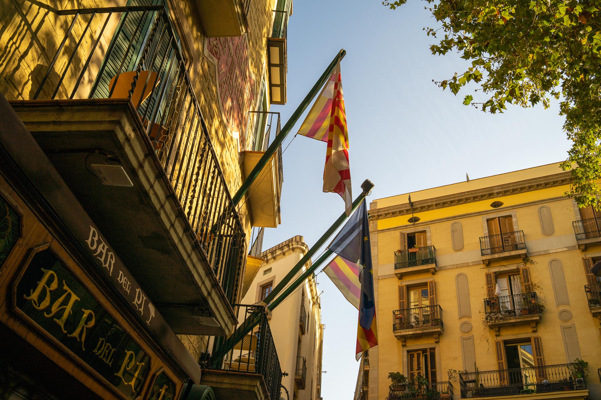 The painted glass sign of Bar del Pi in Barcelona seen from below in low golden light, beneath an iron balcony with green shutters, with the striped and crossed flag of the city and the Spanish and European flags on a pole, and a yellow-rendered building with wooden shutters opposite.