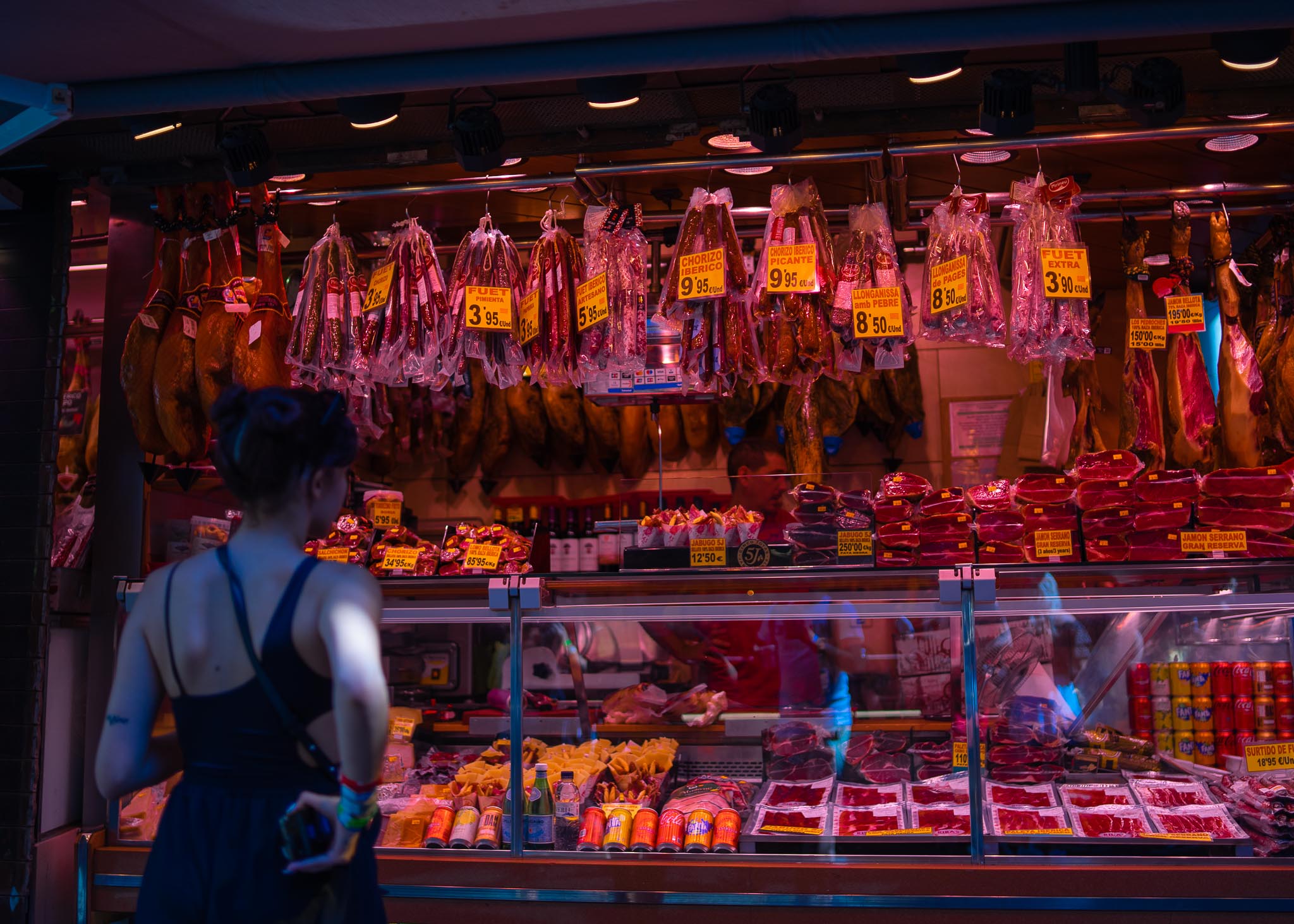 A charcuterie stall in La Boqueria in Barcelona under pink and orange light: whole cured hams hanging in rows, bagged fuet, chorizo and llonganissa strung across the front with large orange handwritten price tags, a chilled counter of sliced meats and cheeses below, and a woman with her back to the camera standing at the glass.