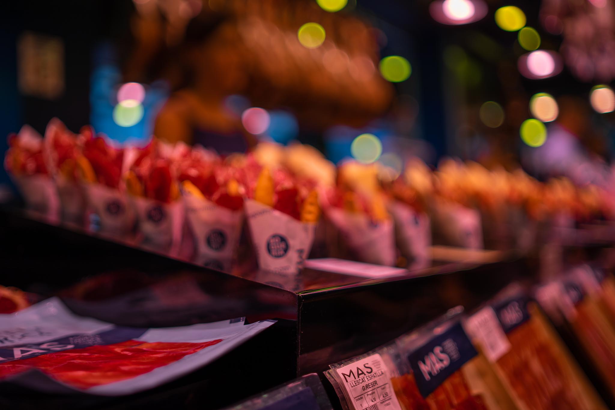 A row of paper cones filled with slices of cured ham in a lit market display case, photographed at a shallow angle so the further cones dissolve into coloured bokeh, with vacuum-packed sliced ham and a printed price label on the counter in the foreground.