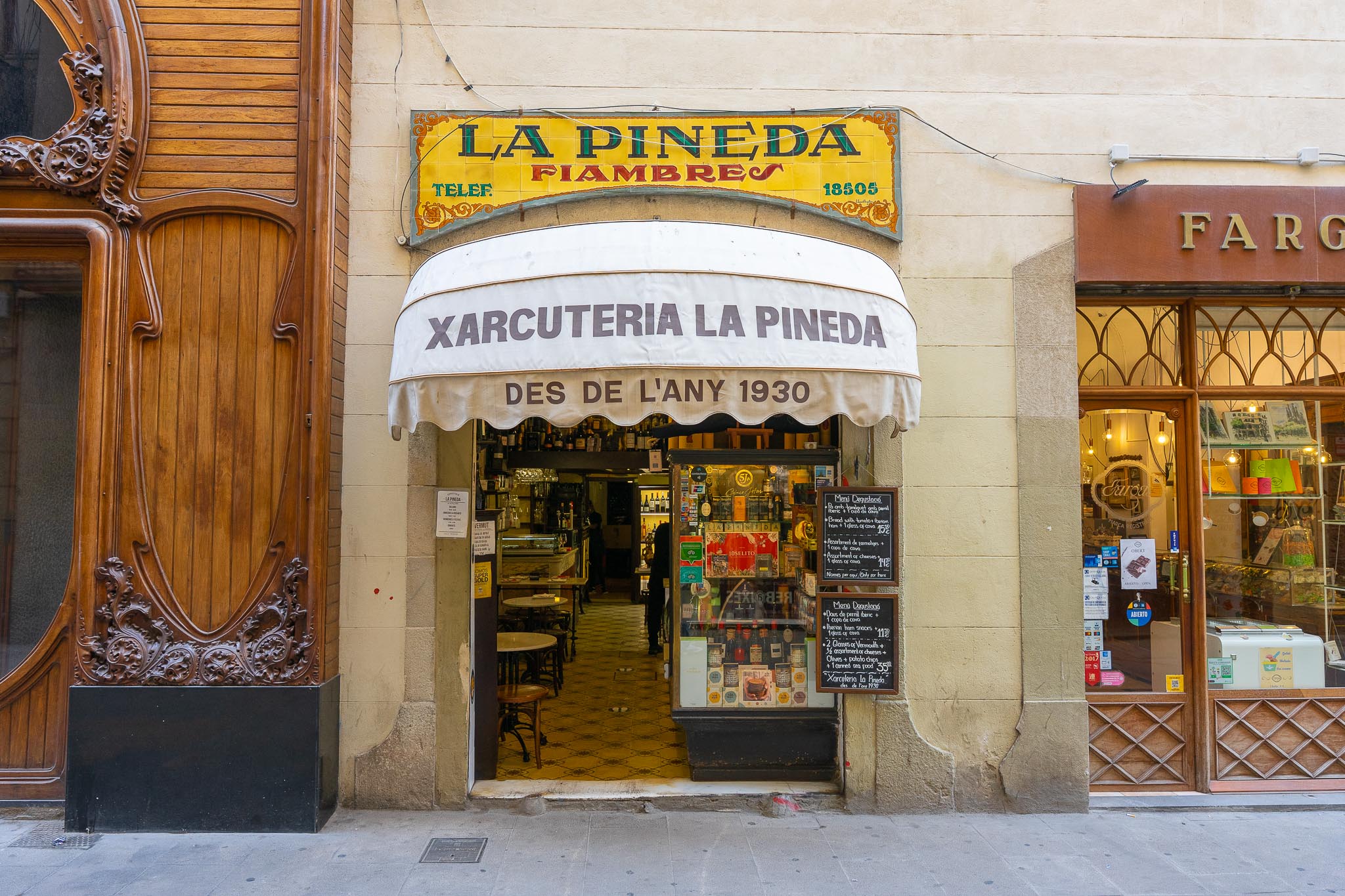 The shopfront of Xarcuteria La Pineda on Carrer del Pi in Barcelona: a glazed tile sign reading LA PINEDA FIAMBRES above a white awning lettered XARCUTERIA LA PINEDA and DES DE L'ANY 1930, an open doorway showing small marble-topped tables and a lit interior, a window stacked with tinned conserves, and two chalkboards listing tasting menus beside the door.