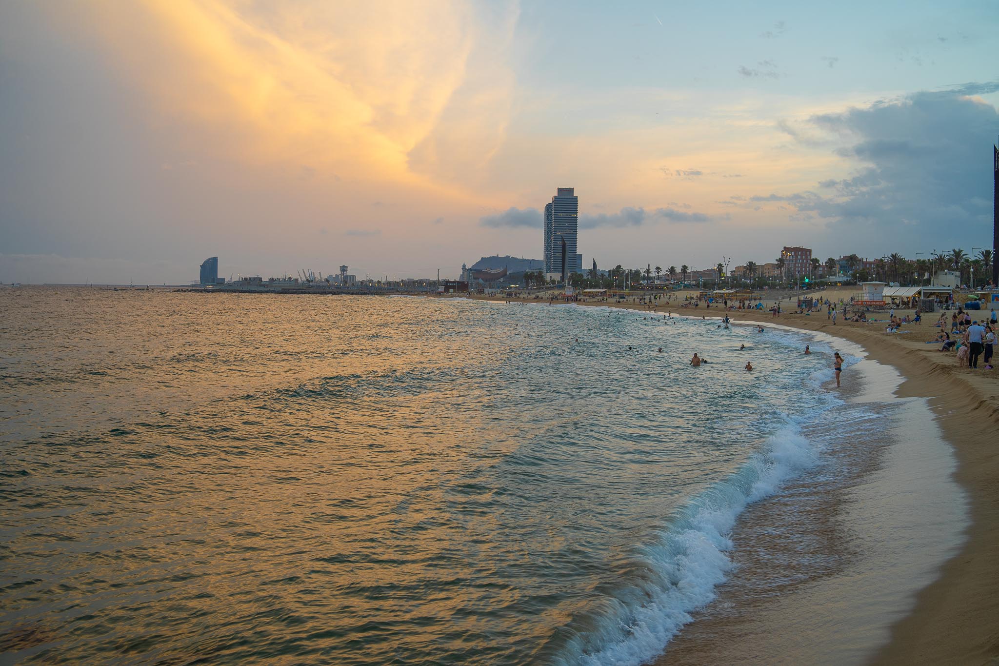 A Barcelona city beach at sunset, low waves breaking on wet sand in the foreground and curving away to the right, a scatter of swimmers and bathers along the shoreline, and the tall tower of Port Olímpic with the low green hill of Montjuïc and a sail-shaped hotel on the horizon under a gold and grey sky.