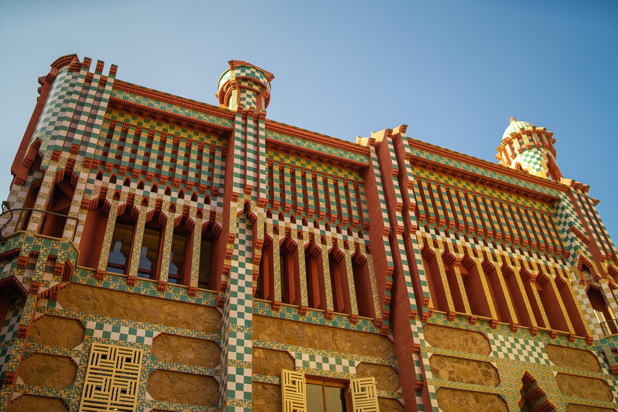 The upper storeys of Casa Vicens in Gràcia, Barcelona, seen from below against a clear sky: rough stone walls banded with green-and-white chequered tiles and rows of orange-and-yellow floral tiles, projecting brick corbels, and two turrets capped in green chequerwork.