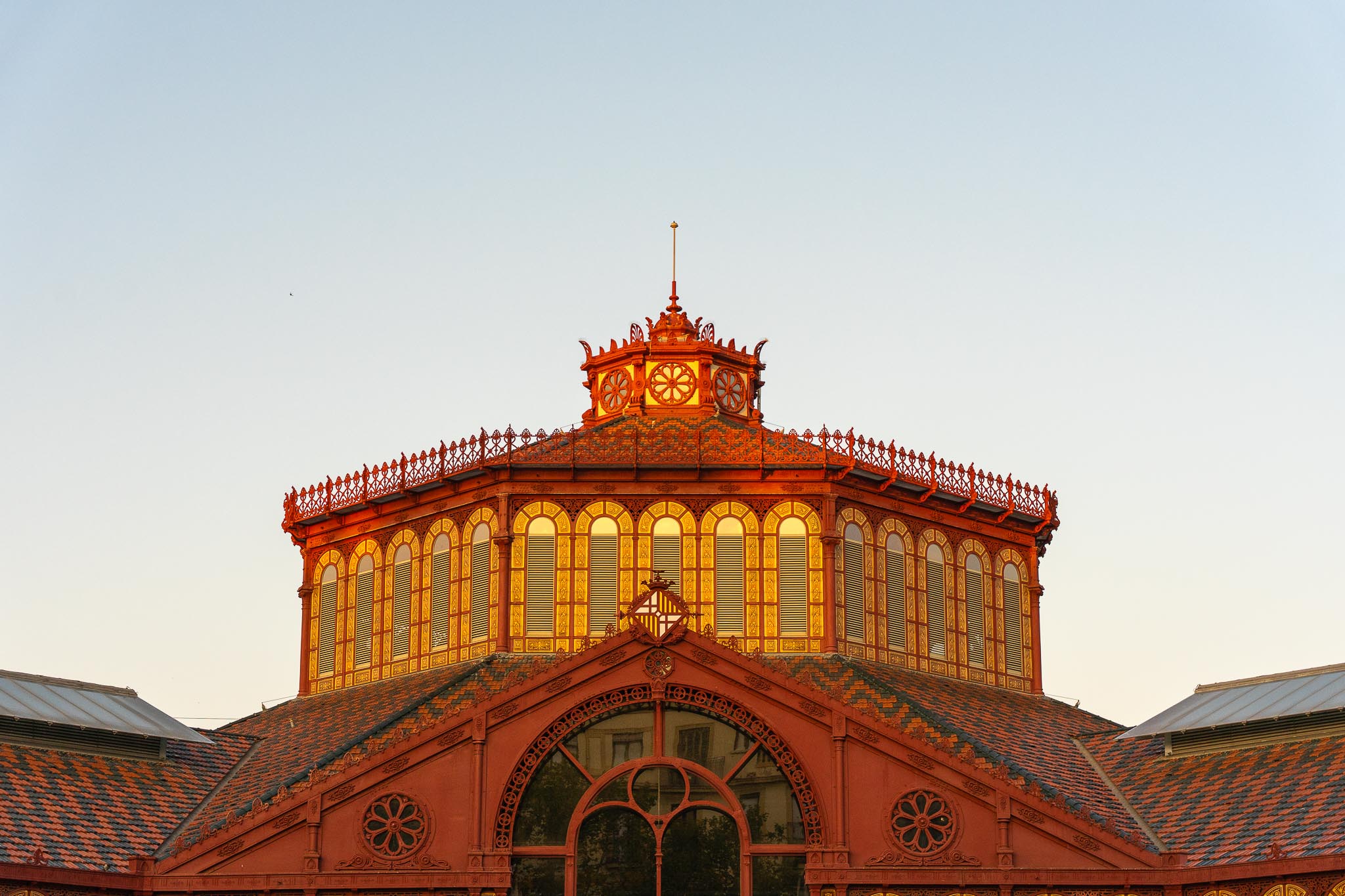 The roof of the Mercat de Sant Antoni in Barcelona at sunset: a red-painted iron octagonal lantern with tall arched shuttered windows glowing gold, standing above a patterned glazed-tile roof and a gable carrying the Barcelona coat of arms, against a pale sky.