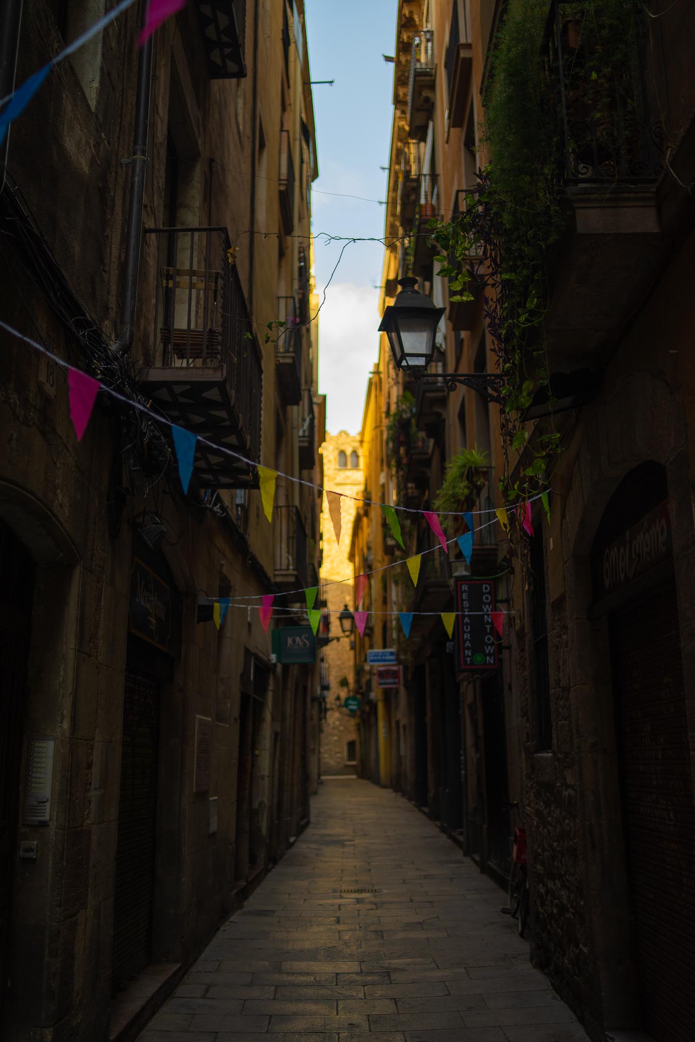 A narrow paved lane in Barcelona's old city, barely wide enough for two people, walled in tall stone buildings with iron balconies and trailing plants, strings of pink, blue, green and yellow bunting overhead, a restaurant sign projecting from the right, and a medieval stone tower catching gold light at the far end.