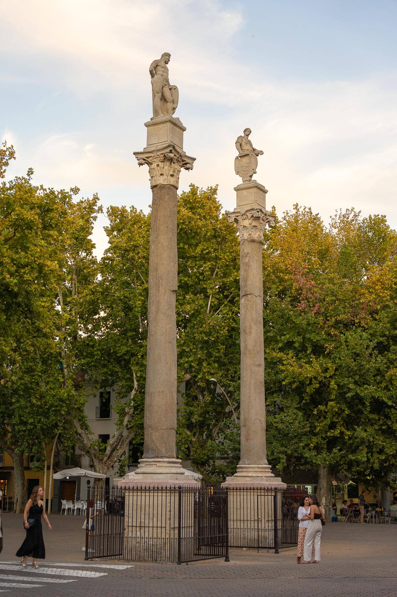 Two tall Roman columns inside iron railings at the southern end of the Alameda de Hércules in Seville, carrying statues of Hercules and Julius Caesar, with plane trees turning yellow behind them, café tables and a few passers-by in evening light.