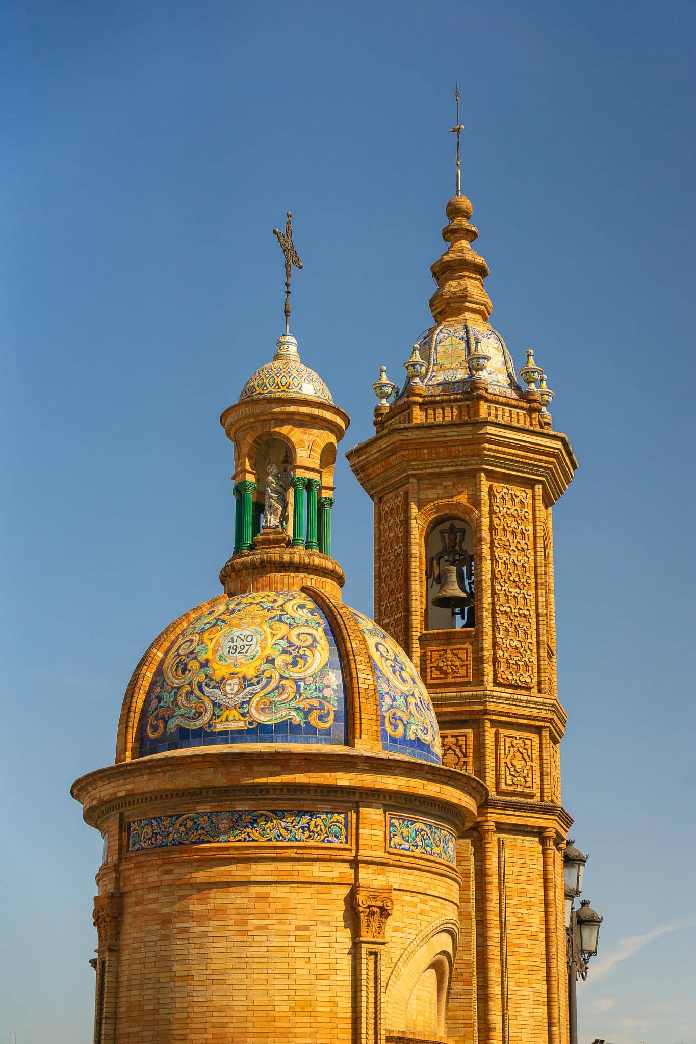 The Capilla del Carmen at the Triana end of the Puente de Isabel II in Seville: a brick drum topped by a glazed ceramic dome bearing the date AÑO 1927 in a cartouche, beside a taller octagonal brick bell tower with tiled cupola and a single bell.