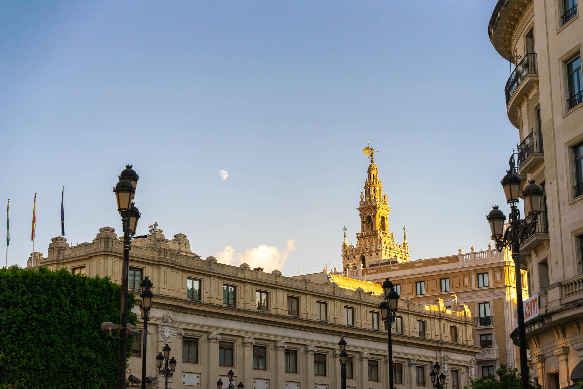 The upper stage of the Giralda catching low gold light above the roofline of a large stone civic building in central Seville, with three flagpoles, ornate iron street lamps and a half moon in a pale sky.