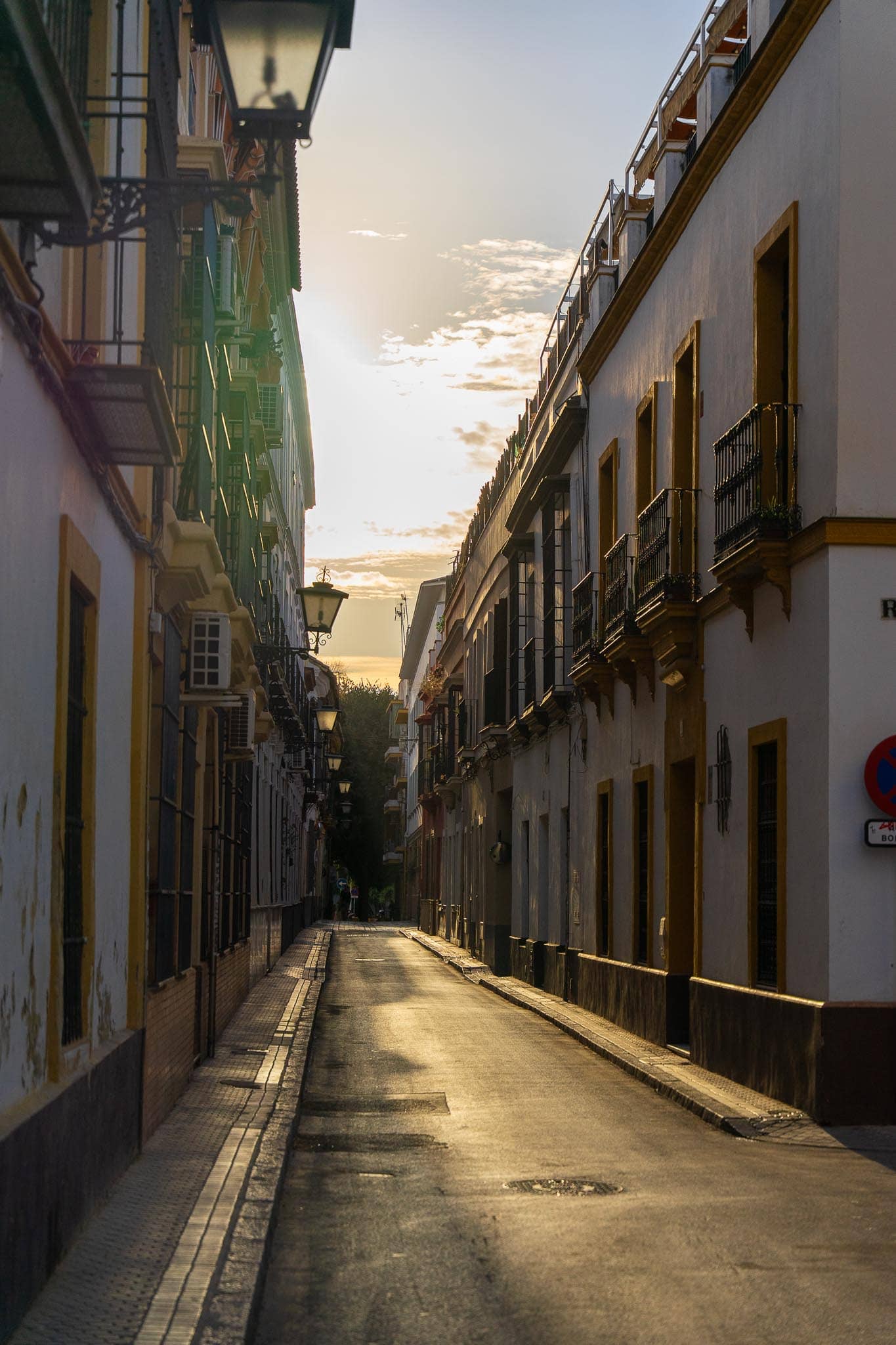 A narrow, empty residential street in Seville with the low morning sun coming down its length: whitewashed and ochre houses with wrought-iron balconies and hanging lanterns on both sides, a single narrow pavement, and trees at the far end.