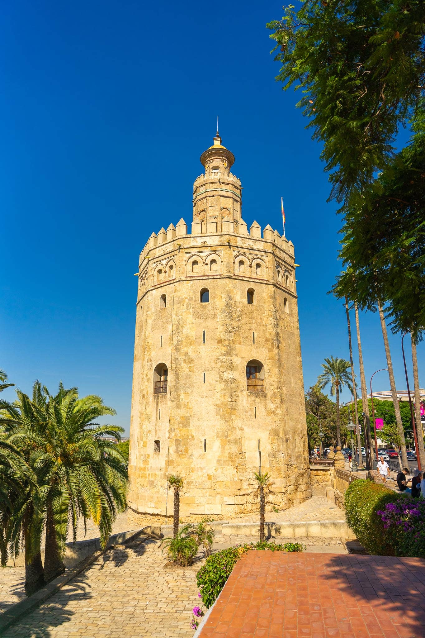The Torre del Oro seen from the landward side among tall palms, its battlemented drum and golden-tiled turret against a deep blue sky, with a Spanish flag flying and brick paving in the foreground.