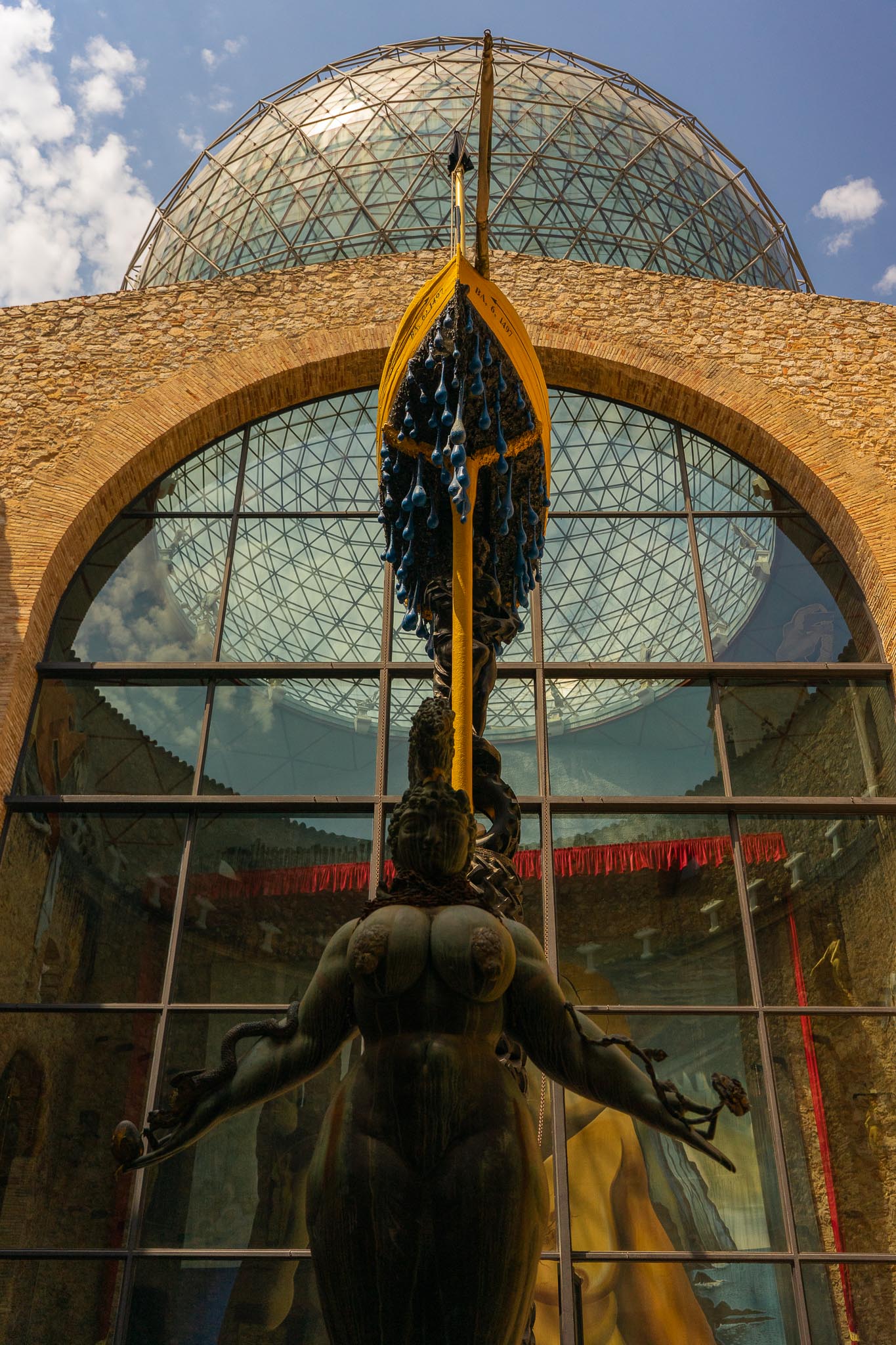 Looking up at the Dalí Theatre-Museum in Figueres: a large geodesic glass dome on a rough stone drum above a broad brick arch, and in the foreground a dark bronze female figure seen from behind holding a long gilded oar, with a cluster of blue teardrop shapes hanging beside it.