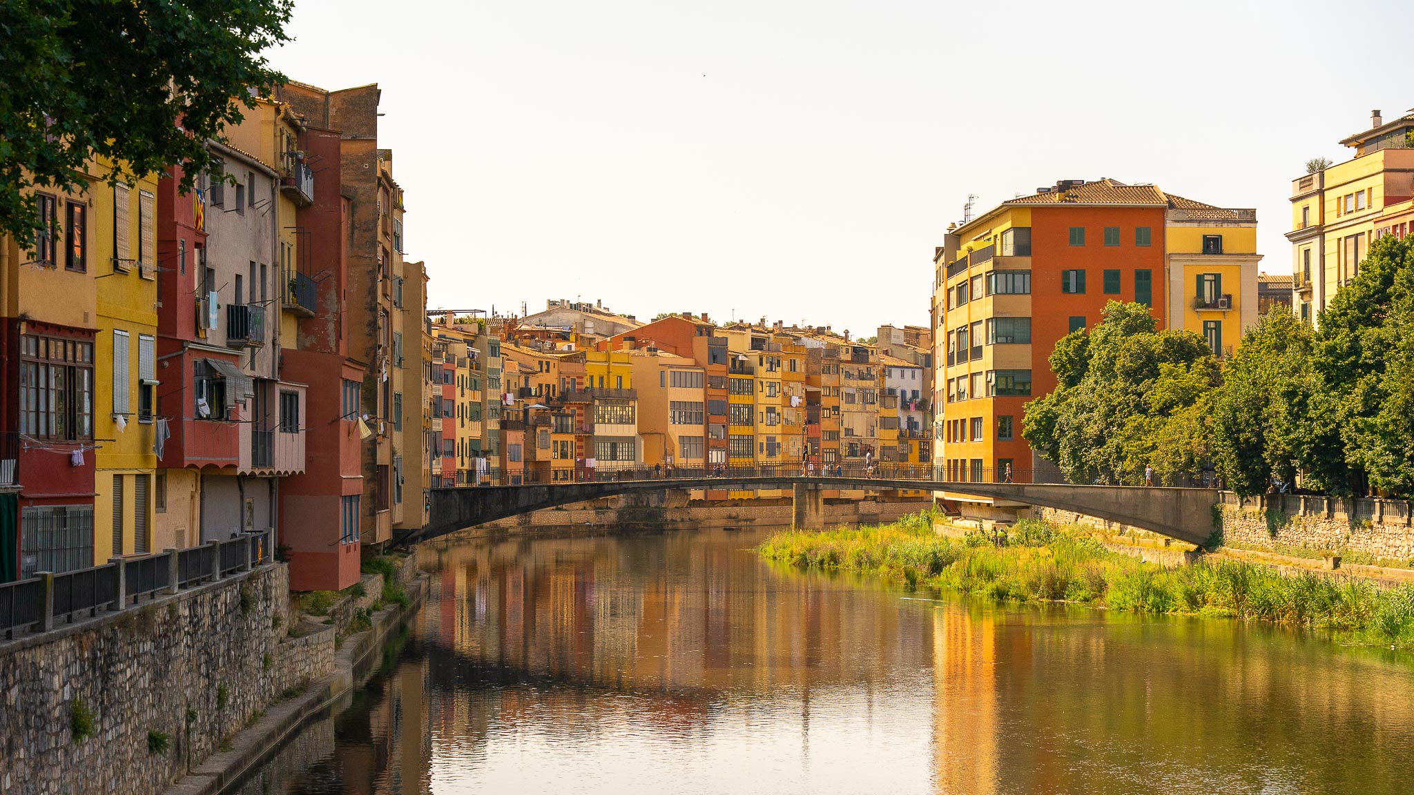 The painted houses of Girona rising straight out of the Onyar in low evening light — ochre, red and yellow façades with shuttered windows, washing on balconies and a Catalan flag — with a low pedestrian bridge crossing the river, reeds on a mid-stream bank, and the whole scene reflected in still brown water.
