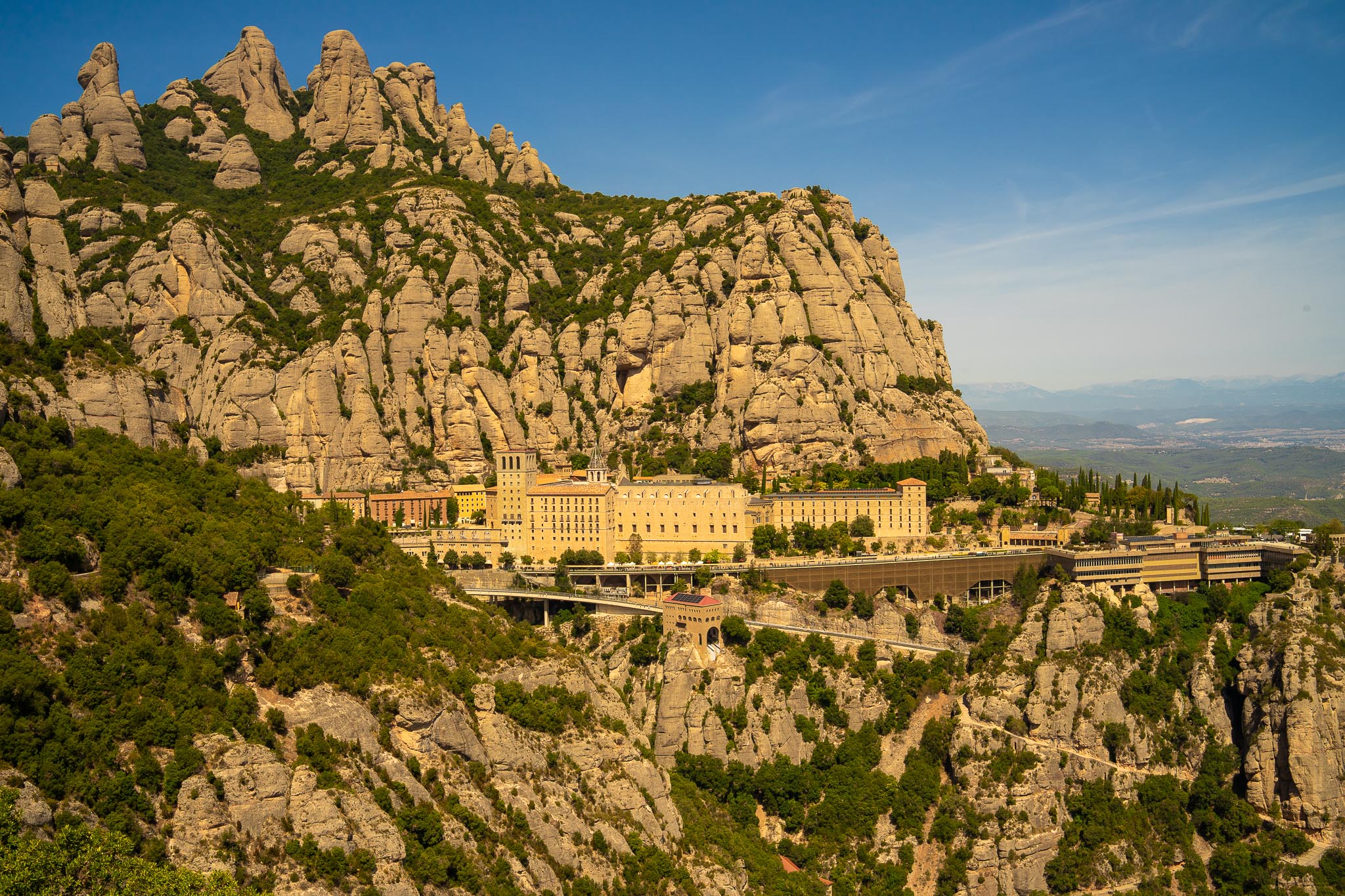 The monastery of Montserrat on its ledge beneath the mountain's serrated conglomerate pinnacles: a long range of pale stone and ochre buildings with a bell tower, terraces and a viaduct built out over the drop, dark green pines above and below, and the hazy plain and distant hills to the right under a deep blue sky.