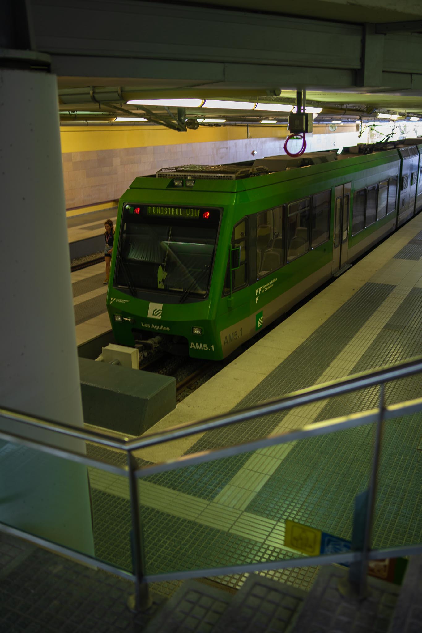 A green rack-railway train standing at a covered platform, its destination board reading MONISTROL VILA, with Ferrocarrils de la Generalitat markings and the unit name Les Agulles on the nose, seen from above across a metal railing, with a single person waiting further down the platform.