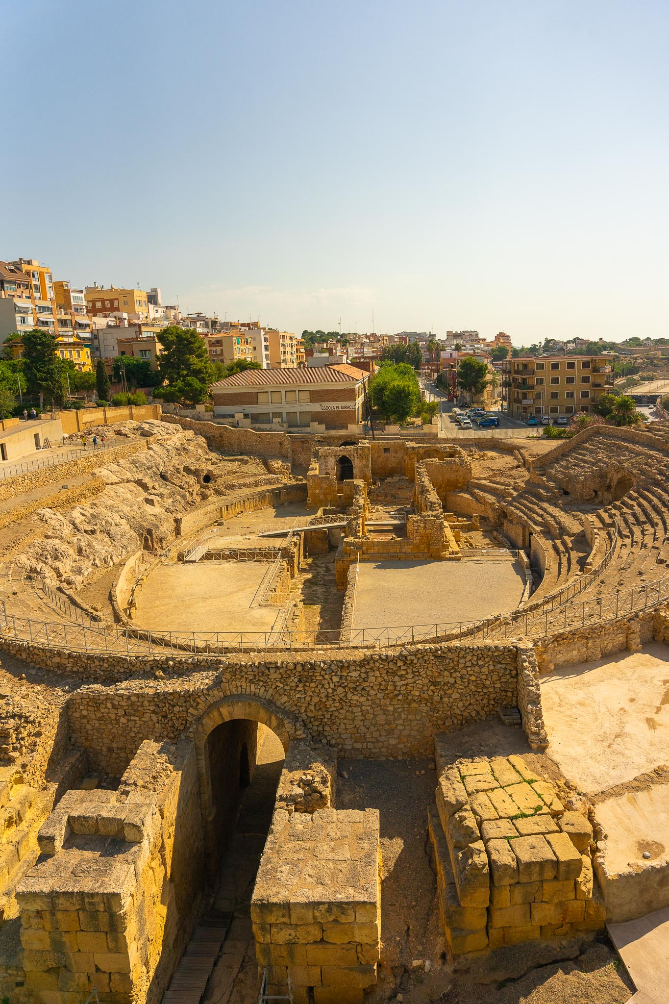The Roman amphitheatre at Tarragona seen from above: the elliptical arena with the stone footings of a later church standing in it, surviving tiers of seating cut into the rock on the right, a vaulted stone passage in the foreground, and modern apartment blocks and a school on the slope beyond.