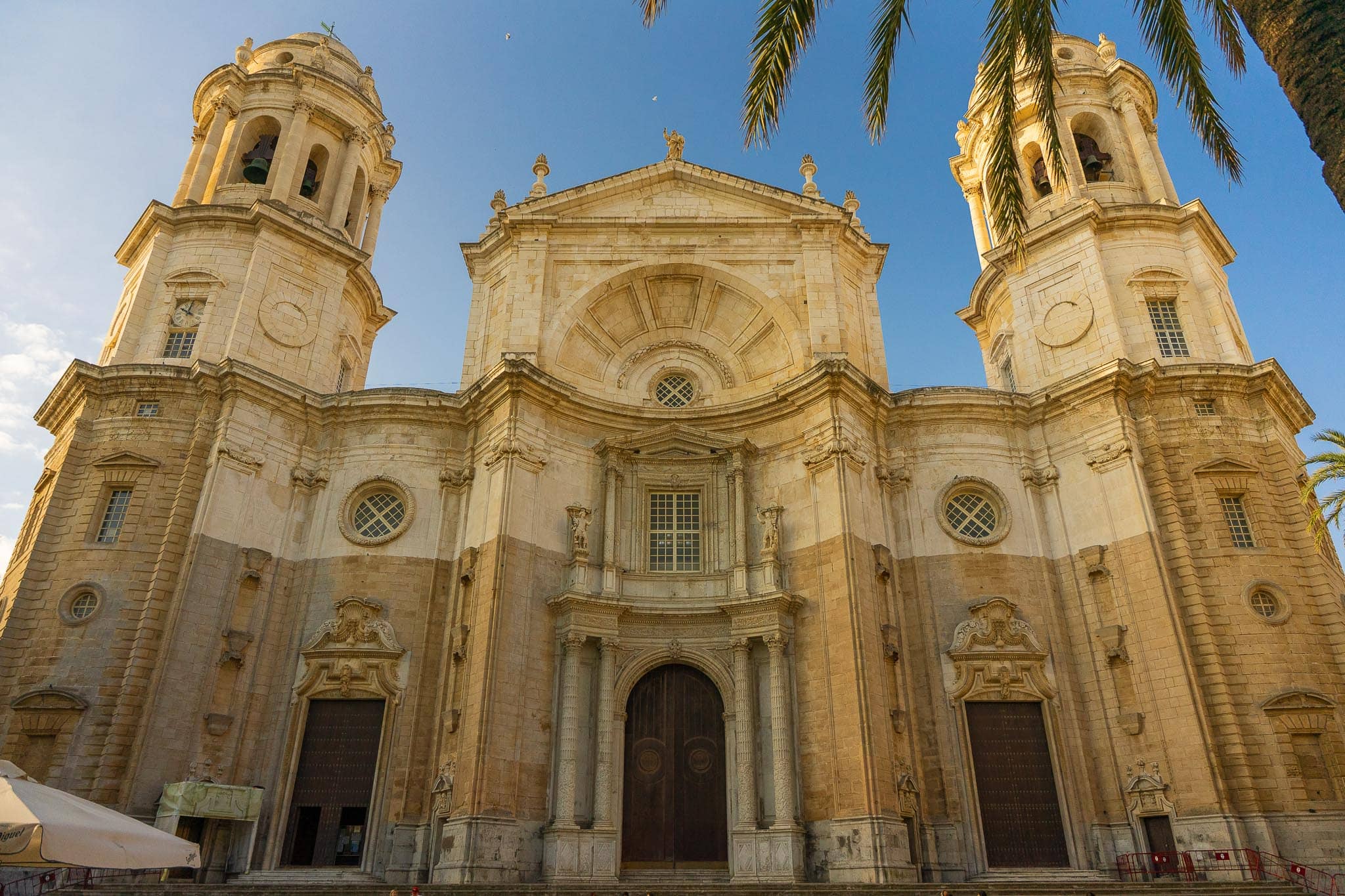 The west front of Cádiz Cathedral: a broad pale limestone baroque façade with two domed bell towers, a large arched central bay and a carved doorway, half of it in shadow and half in golden morning light, with palm fronds crossing the top of the frame.