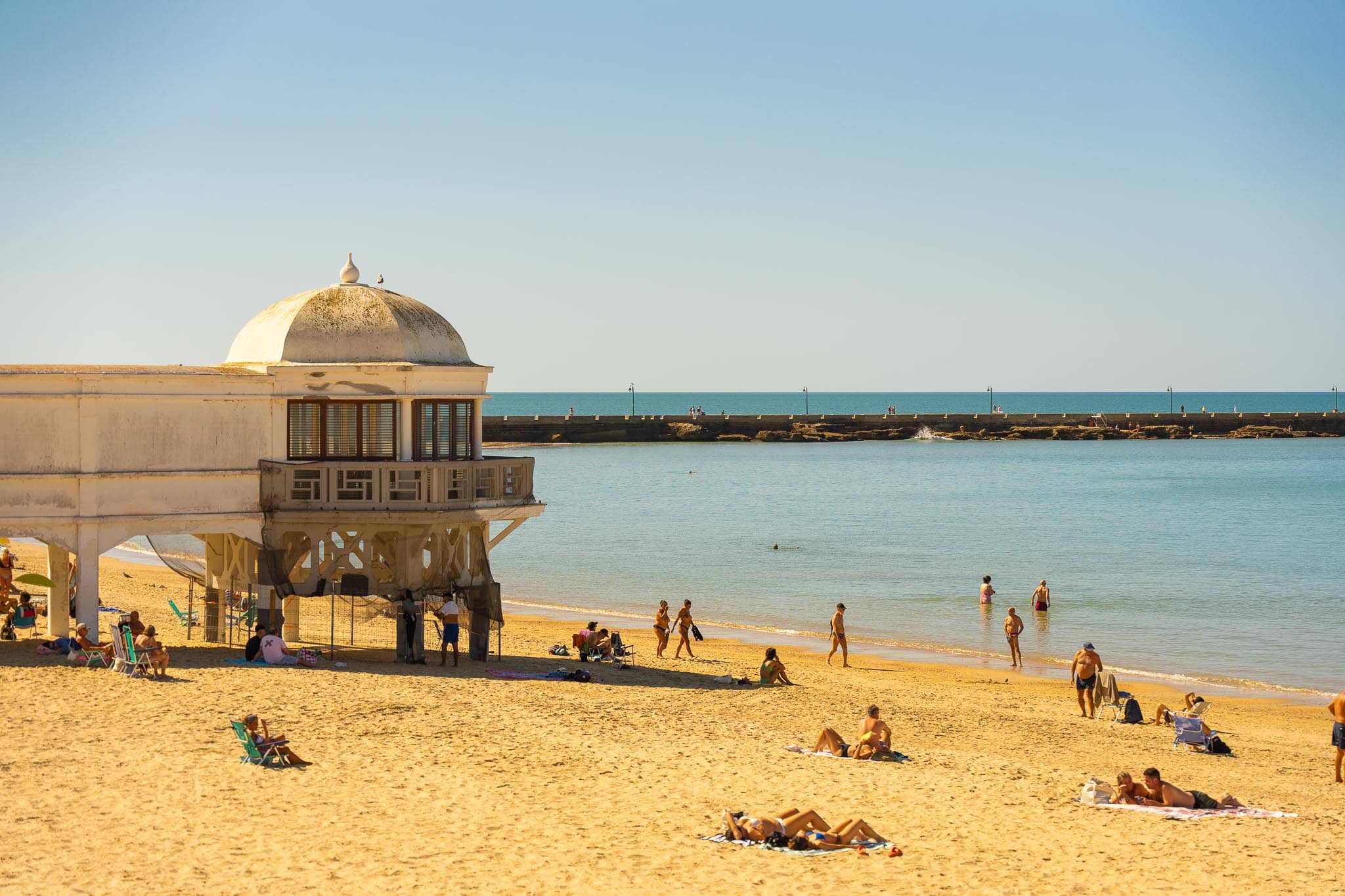 La Caleta beach in Cádiz: a domed white balneario building raised on piers at the left, a long stone breakwater running out towards a sea fort, turquoise Atlantic water, and bathers and sunbathers on pale sand.