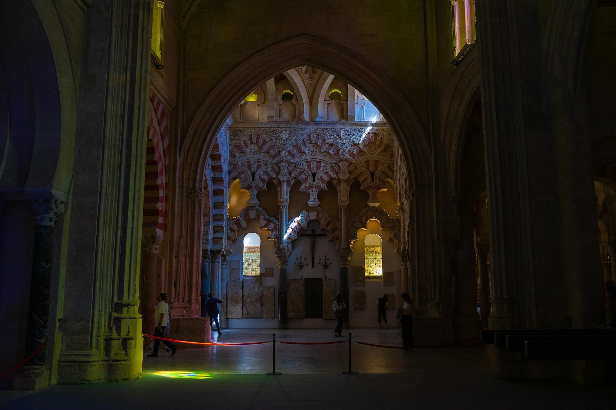 Inside the Mezquita-Catedral of Córdoba: a tall Christian Gothic pointed arch in the foreground framing the interlacing red-and-white polylobed Islamic arches and carved plasterwork of the Villaviciosa chapel beyond, with a crucifix below and coloured light from stained glass thrown across the stone floor.
