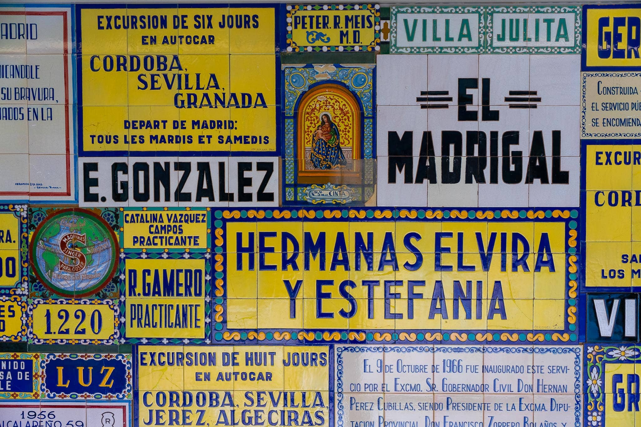A wall of old Andalusian trade azulejos: a large yellow and blue tiled panel advertising a six-day coach excursion to Cordoba, Sevilla and Granada departing Madrid every Tuesday and Saturday, surrounded by tiled shop and house signs including E. Gonzalez, El Madrigal, Villa Julita, Hermanas Elvira y Estefania, a Singer sewing-machine roundel and a painted Virgin medallion.