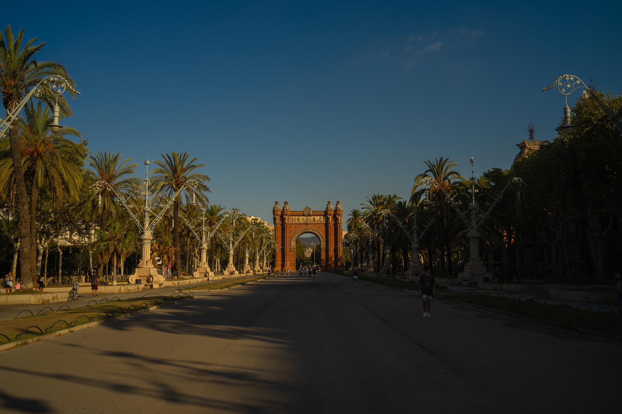 The Arc de Triomf in Barcelona seen down the length of Passeig de Lluís Companys in low evening sun: a red brick arch at the far end of a broad, almost empty roadway, flanked by rows of date palms and tall ornate white iron lamp standards, with a wooded hill visible through the archway.