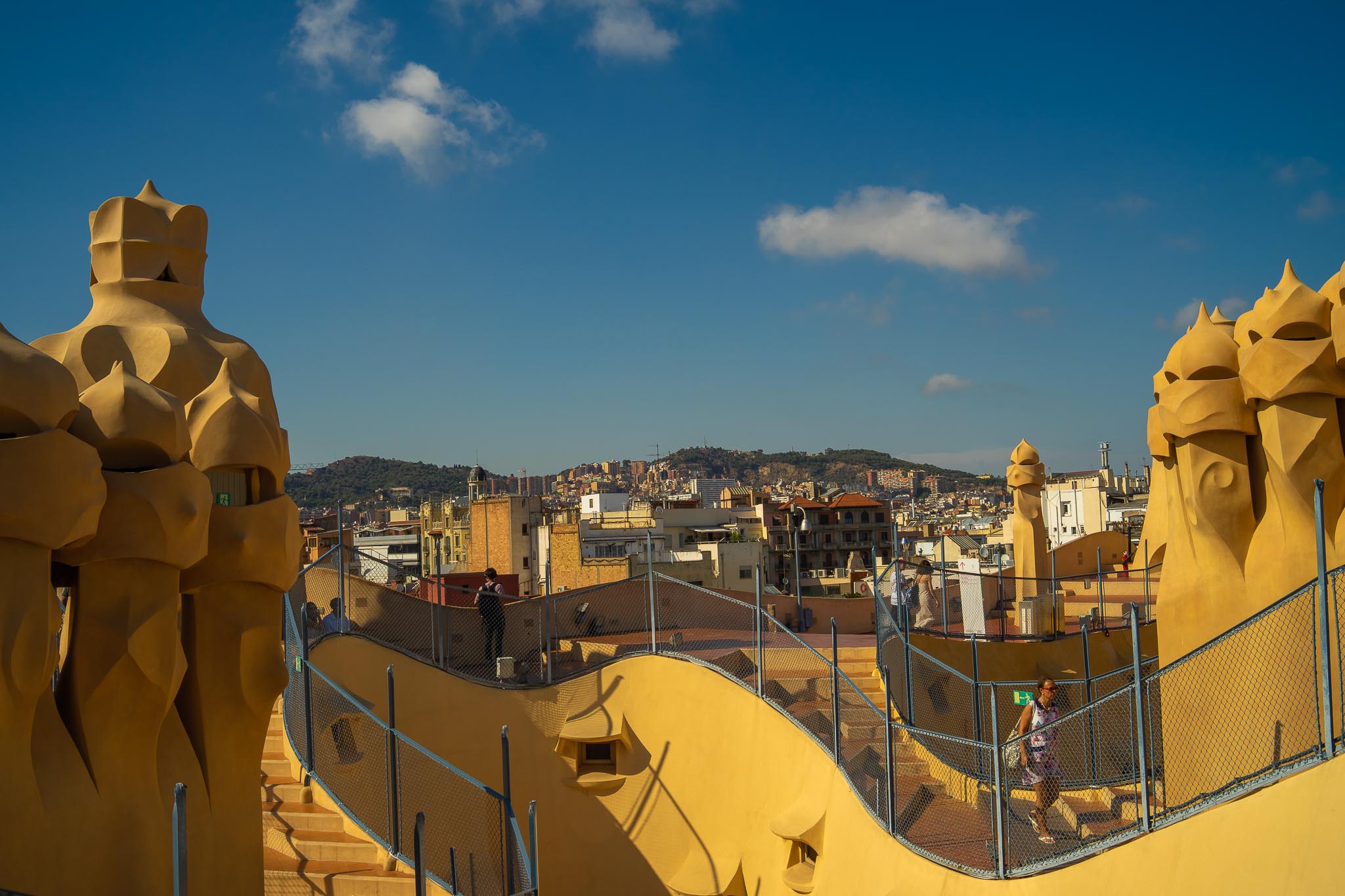 The roof terrace of La Pedrera in Barcelona in low evening sun: ochre rendered chimney stacks with helmeted, faceted heads standing on both sides of a sinuous stepped walkway edged with wire mesh guard fencing, the flat roofs and balconies of the Eixample beyond, and the wooded ridge of Collserola along the horizon under a deep blue sky.