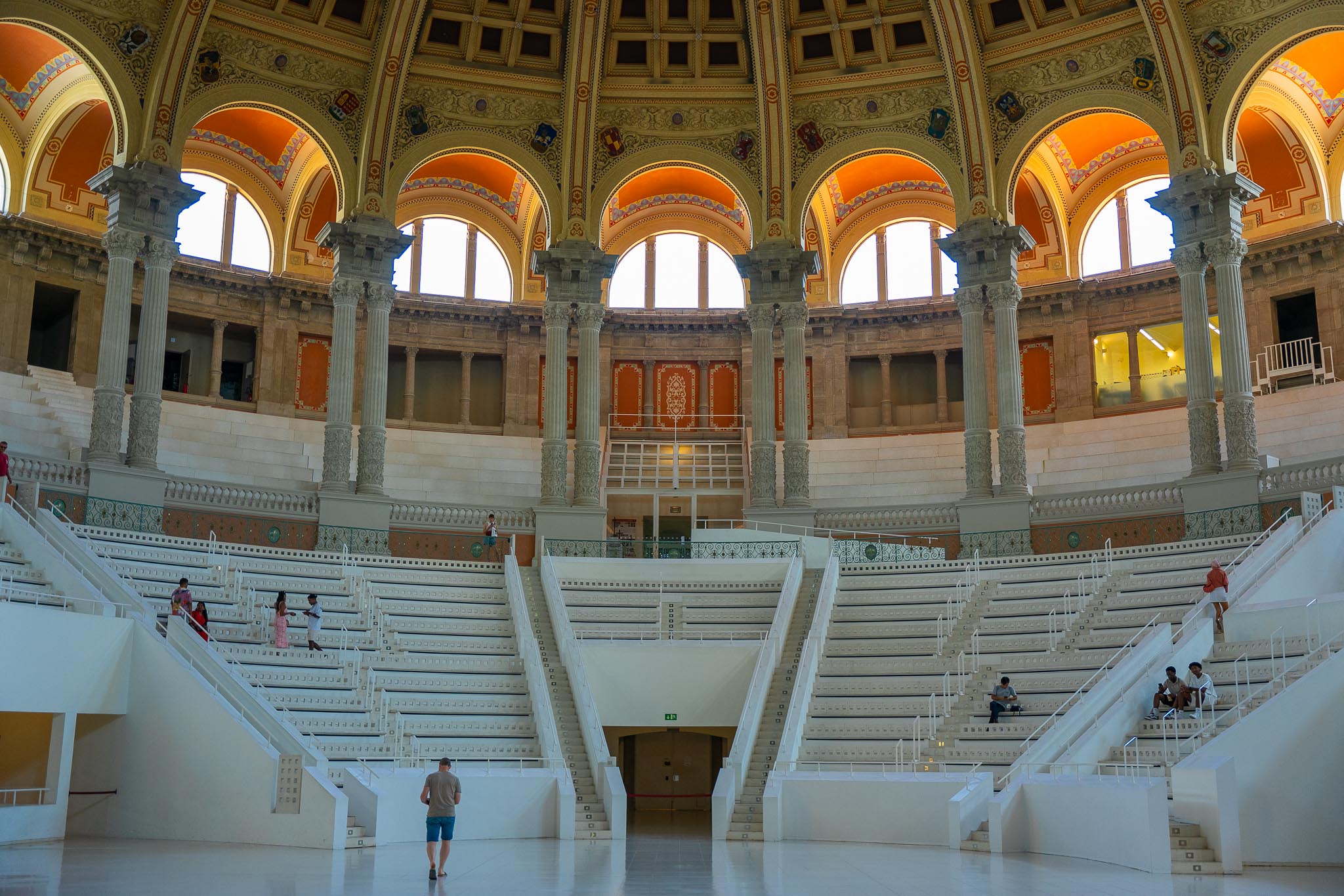 The Oval Hall of the MNAC in Barcelona: tiers of white seating rising on both sides of a broad central staircase beneath a ring of tall Corinthian columns, arched windows filled with white light, and a coffered dome painted in orange, gold and pale blue, with a handful of small figures on the steps and the floor.