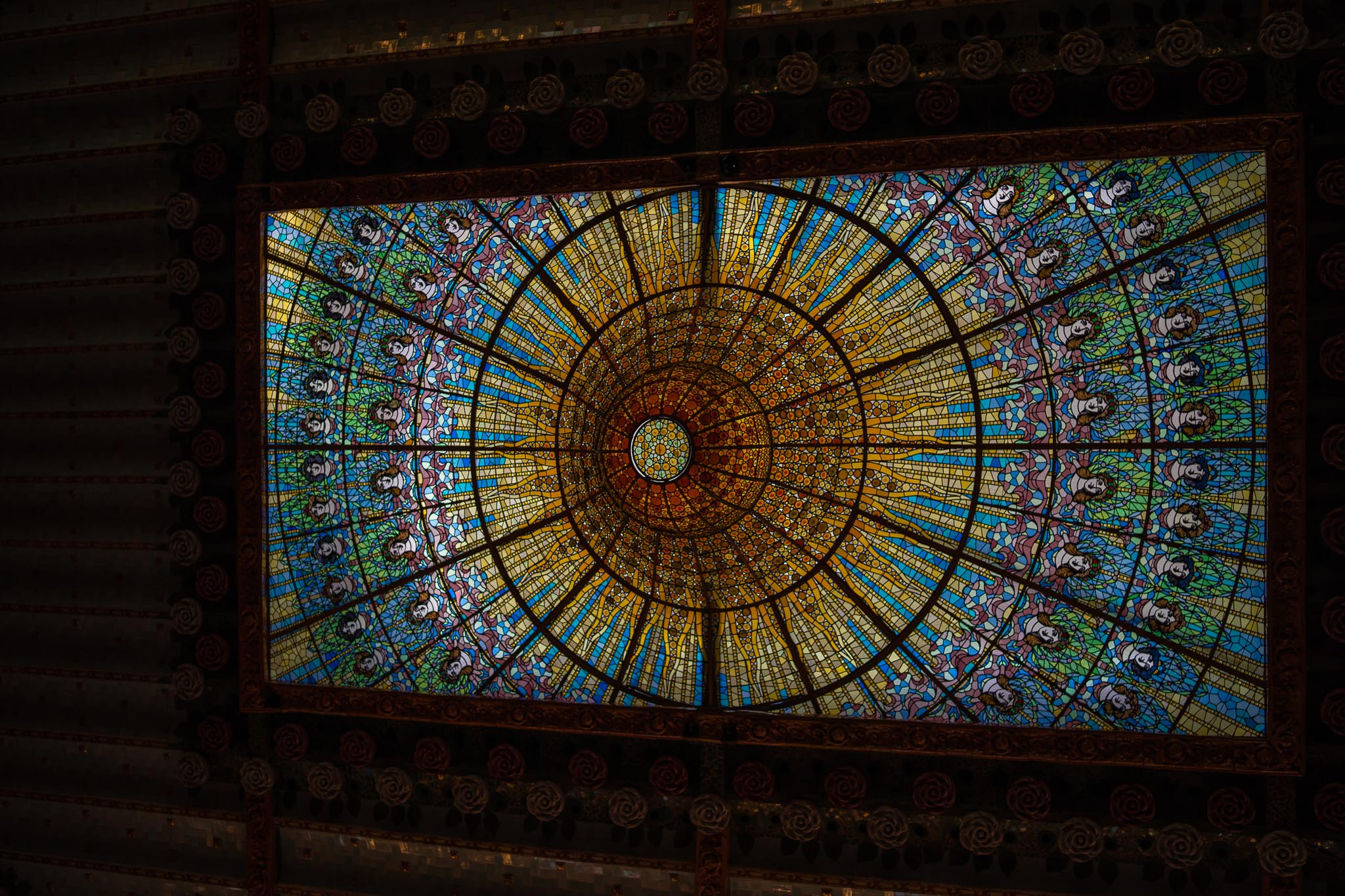 The stained-glass skylight of the Palau de la Música Catalana in Barcelona seen from below: a rectangular frame enclosing an inverted cone of amber and gold glass at its centre, ringed by radiating panes of blue, green and pink glass set with rows of women's faces, all surrounded by the dark carved and rose-patterned soffit of the ceiling.