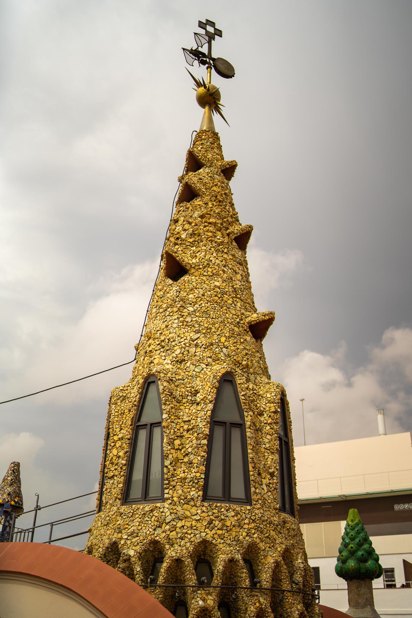The central spire on the roof of Palau Güell in Barcelona seen steeply from below: a tall cone clad in irregular pale limestone fragments, pierced by triangular vents and tall narrow windows, topped by a gilded spiked ball and a wrought-iron weathervane carrying a bat and a four-armed cross, with a green ceramic-scaled chimney to the right and grey cloud behind.