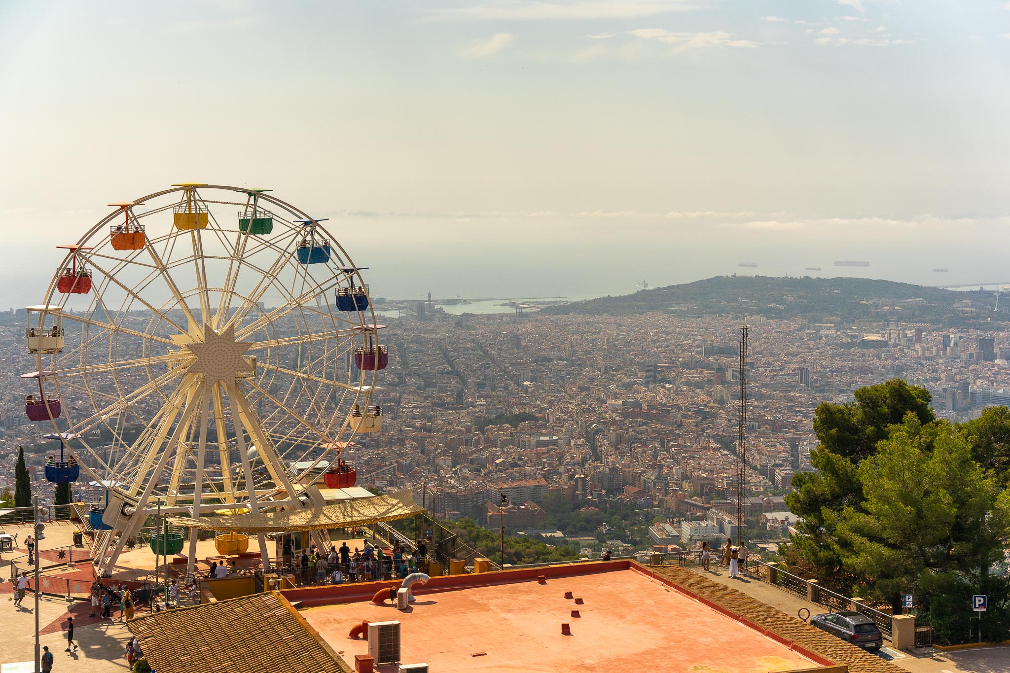 A ferris wheel with brightly coloured open cabins standing on the summit of Tibidabo above Barcelona in hazy afternoon light, the city grid running away below it to the port and the Mediterranean, a lattice mast to the right, a pine in the near foreground and a red-surfaced terrace below.
