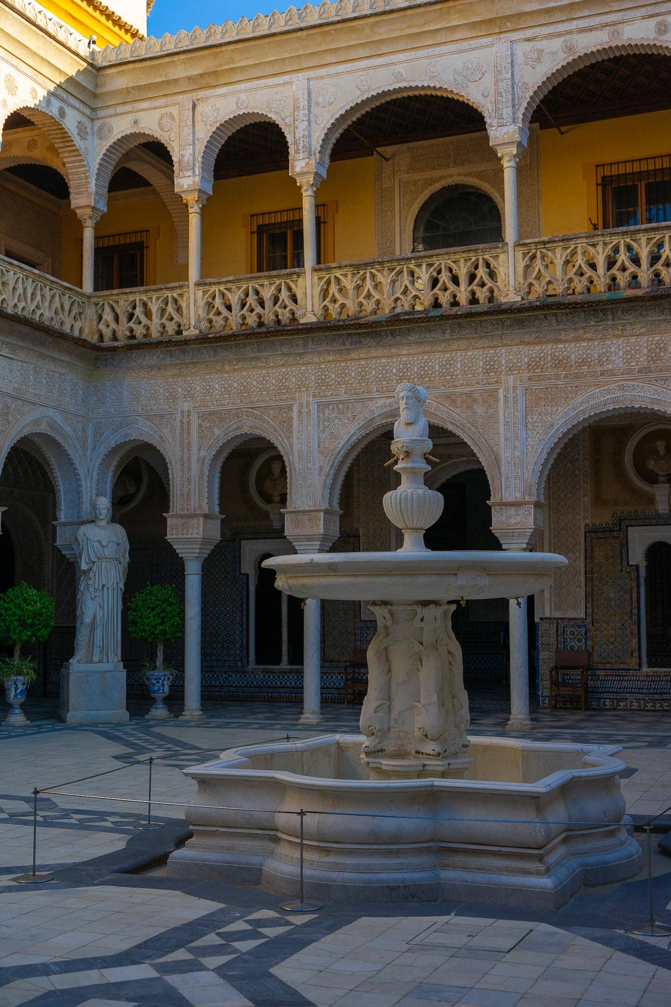 The principal courtyard of Casa de Pilatos in Seville: two storeys of arcading with intricate Mudéjar plasterwork and a Gothic tracery balustrade above, blue and white azulejo dados, Roman busts set in roundels, a standing Roman female statue on a plinth, and a marble fountain with dolphins beneath a bearded Janus head on chequered marble paving.