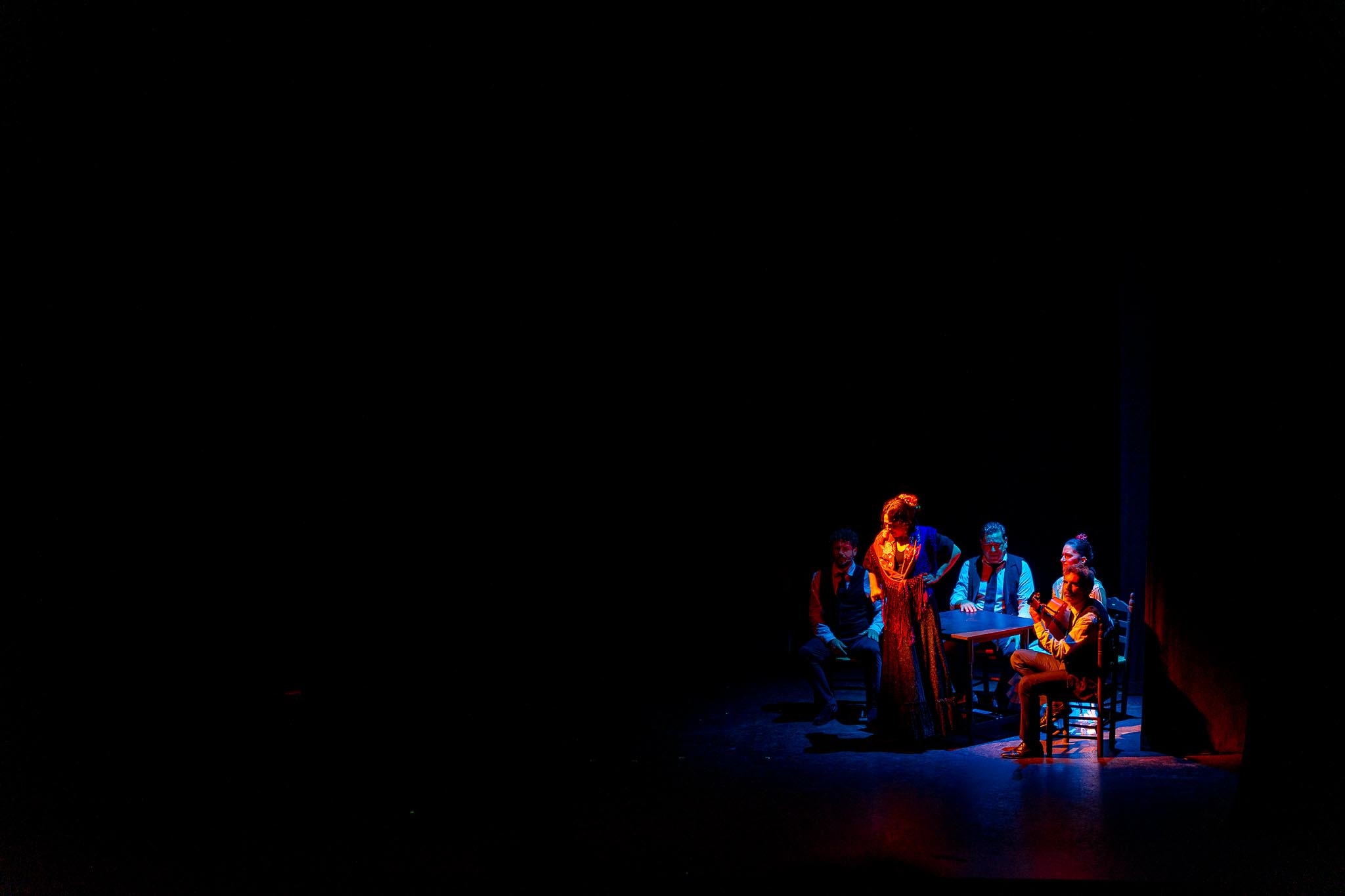 Six flamenco performers on a dark stage lit in blue and red: a guitarist in shadow at the left, a woman standing in a red and gold fringed dress with her hand on her hip, three seated figures around a small table, and a second guitarist playing at the right.