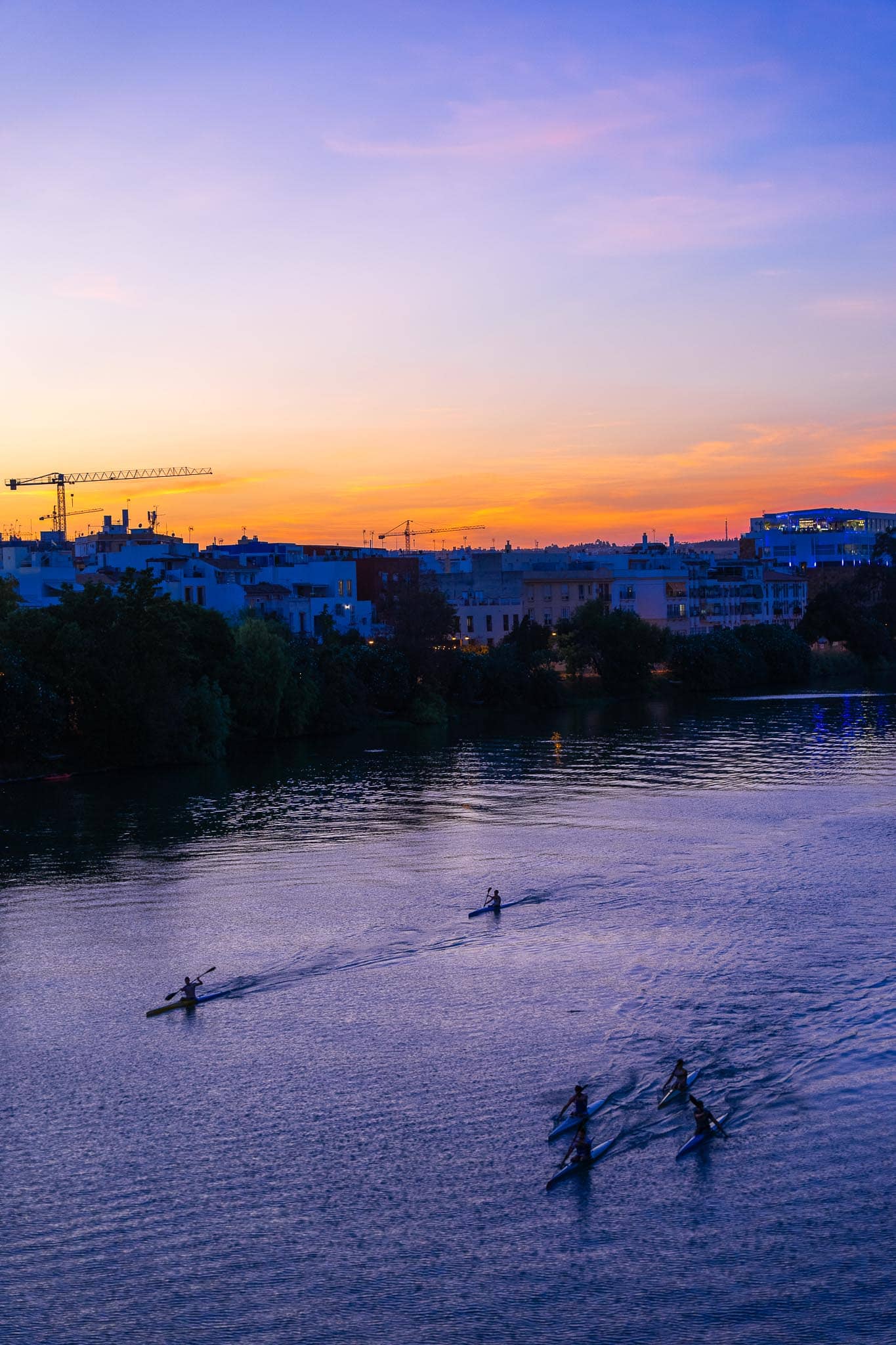 The Guadalquivir at sunset seen from above: a lone kayaker and a group of four paddlers on violet water, the far bank in silhouette with white apartment buildings, trees, construction cranes and a lit modern building, beneath an orange and violet sky.