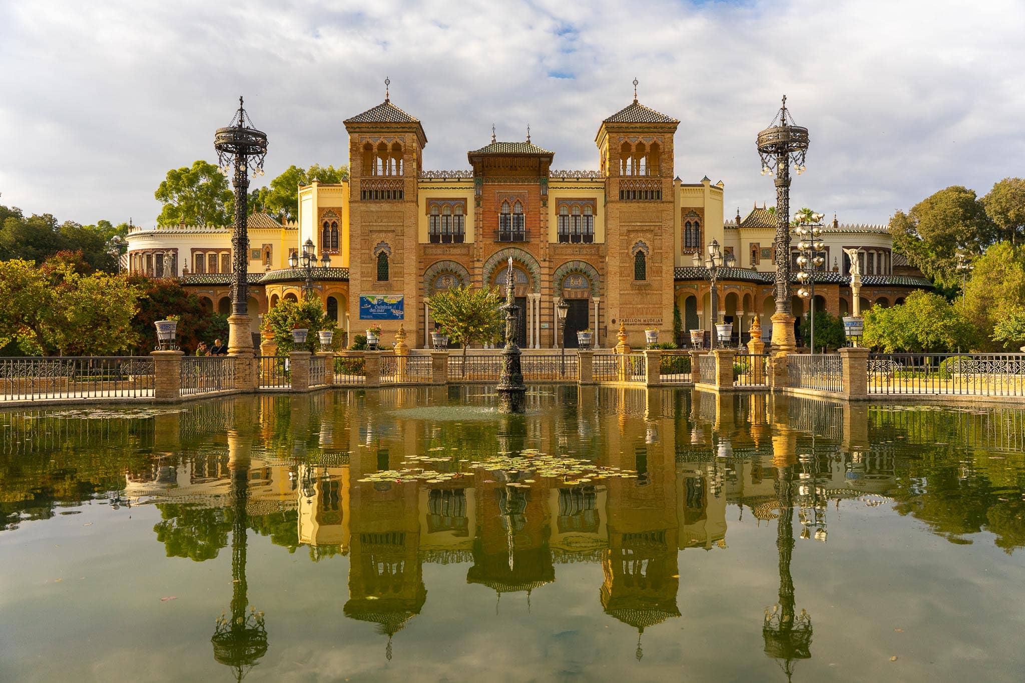 The Pabellón Mudéjar in Parque de María Luisa, Seville, mirrored in its still lily-padded pool: a brick and tile pavilion with two towers and a green-tiled roof, flanked by two tall ornate iron lamp standards, with a small fountain playing in the water.