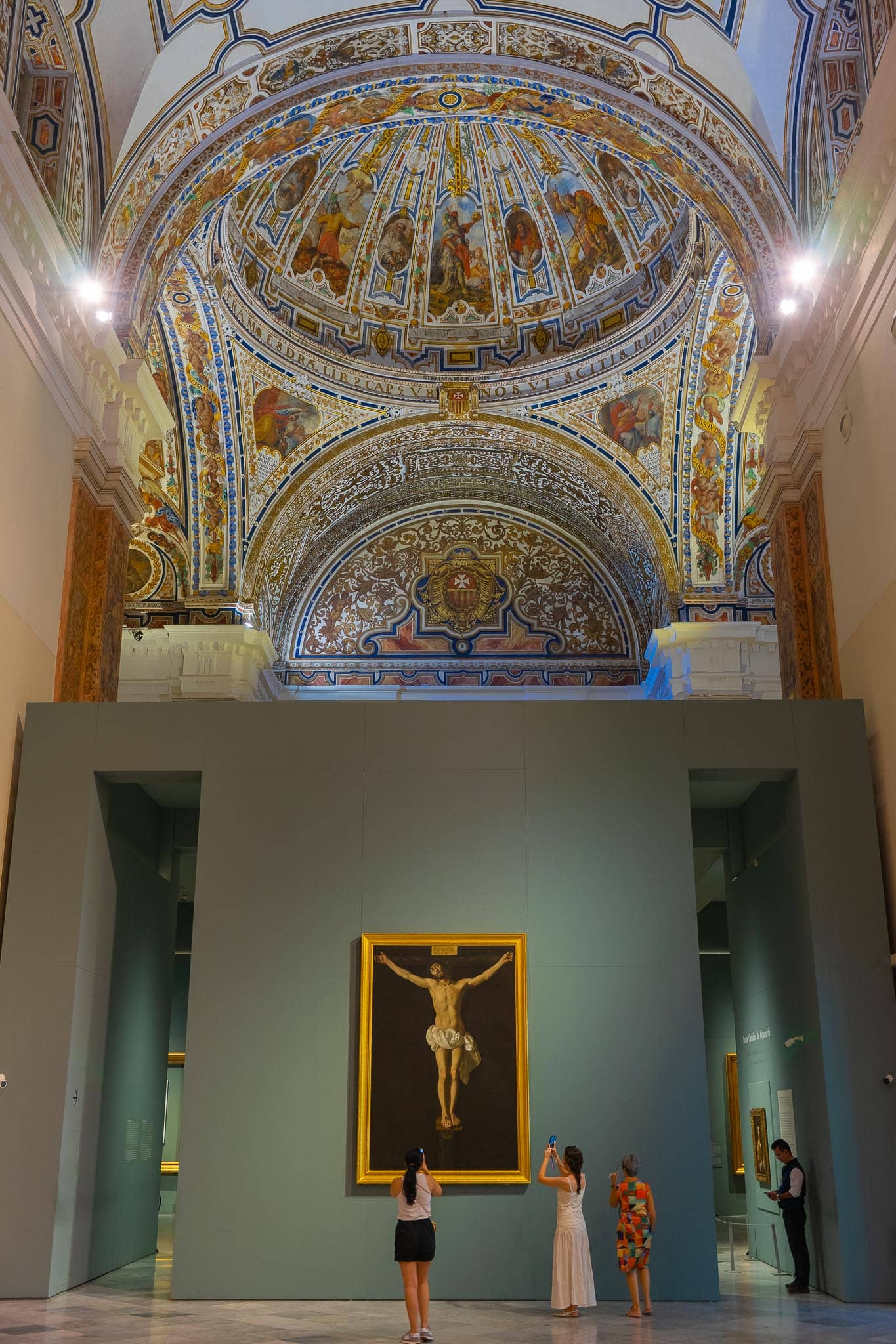 A single large Zurbarán crucifixion in a gilded frame hung alone on a pale partition wall in the Museo de Bellas Artes in Seville, beneath the frescoed dome and painted pendentives of the former convent church, with three visitors photographing it from below.