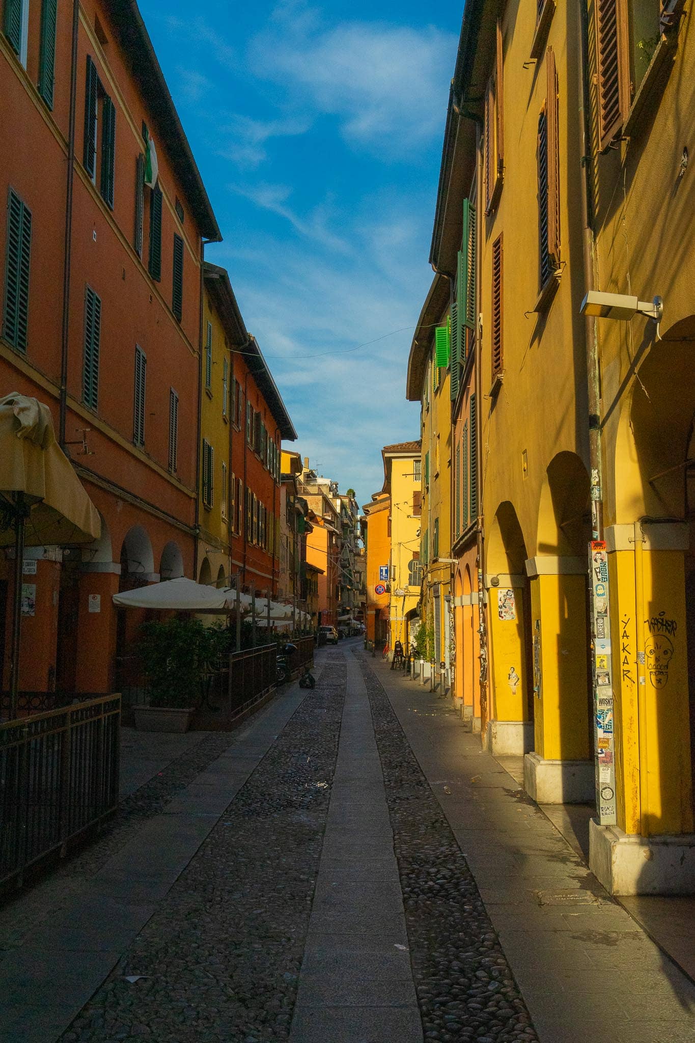 A narrow Bologna street of ochre, yellow and terracotta buildings with green shutters, a strip of blue sky above and cobbled paving running away downhill.
