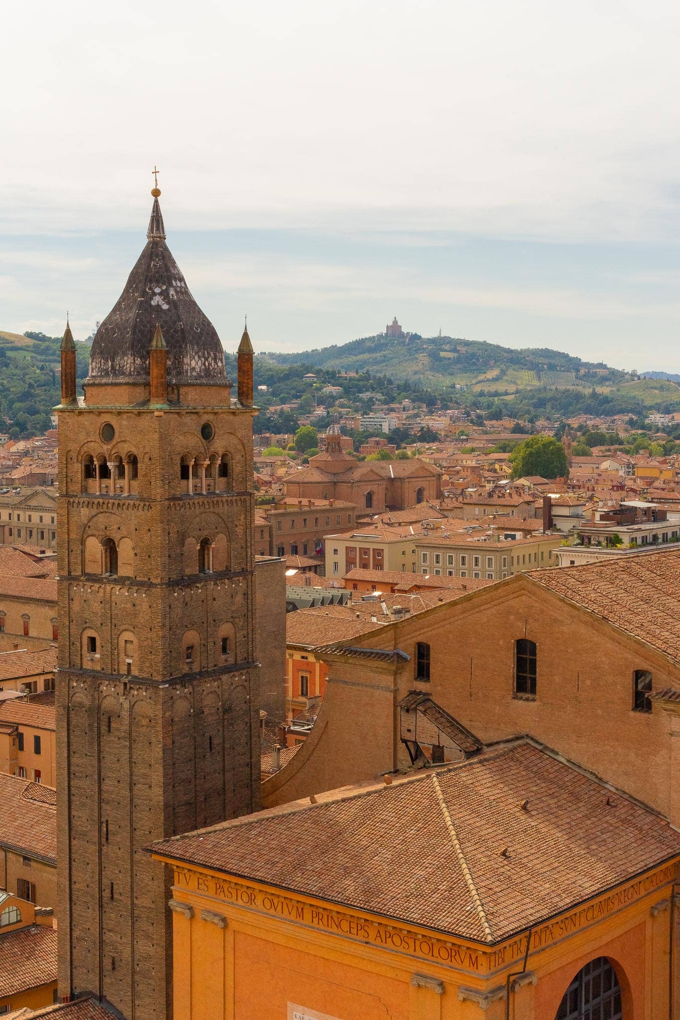 A tall brick and stone campanile rising above the terracotta rooftops of Bologna, with wooded hills and a hilltop sanctuary faint on the skyline.