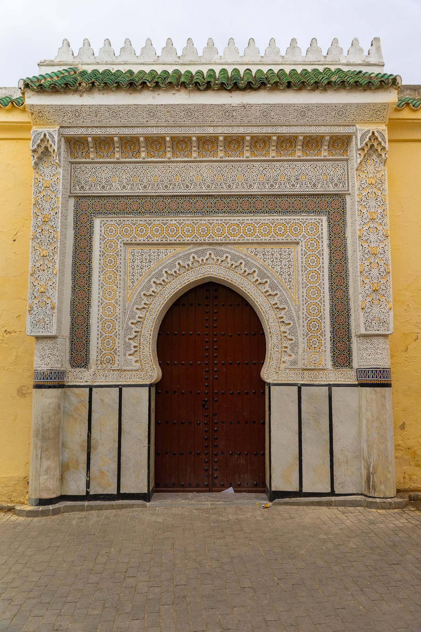 An ornamental gateway in Fez: a cedar door under a horseshoe arch framed by carved white stucco, set in a plain yellow wall beneath a green-tiled cornice.