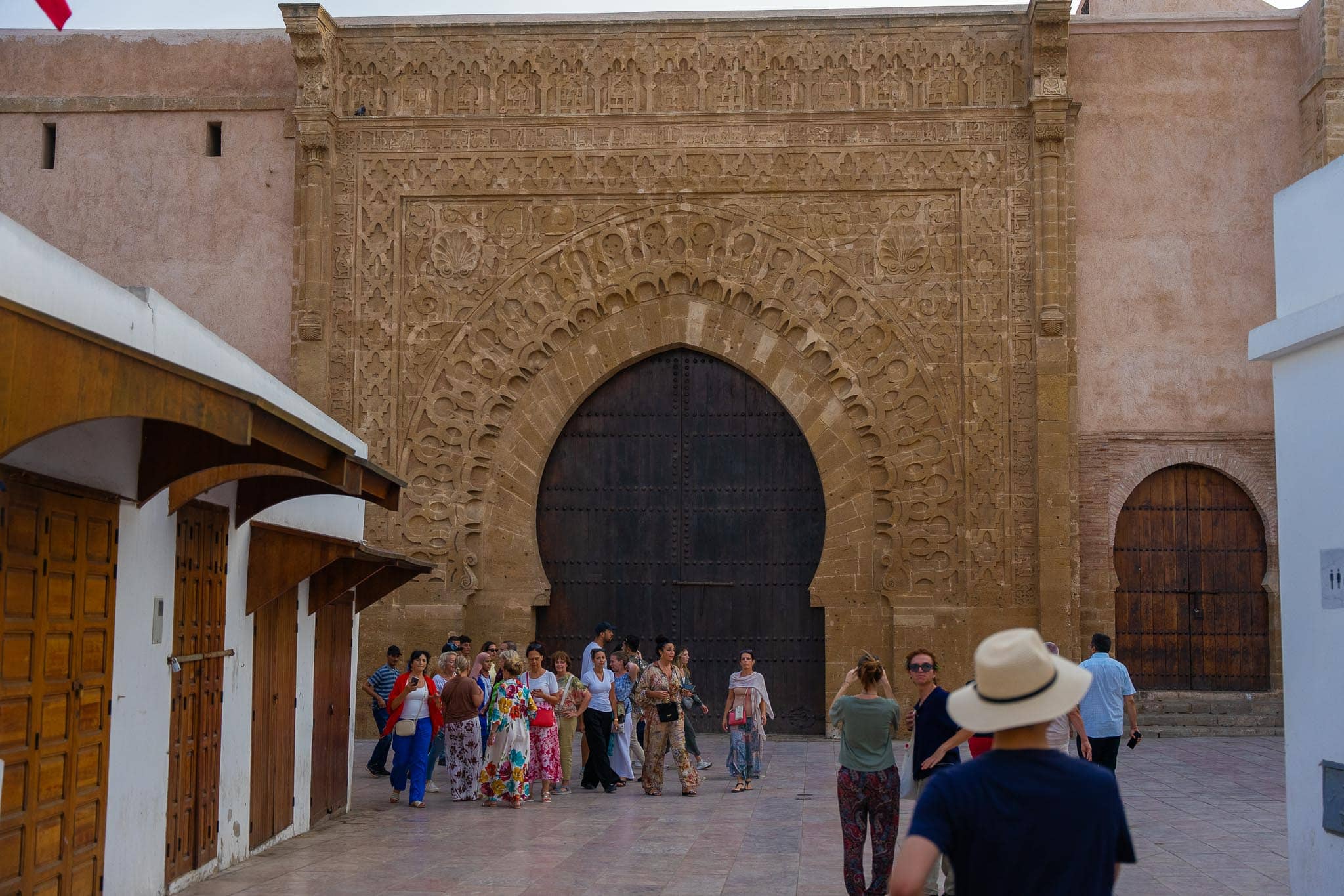 A monumental Almohad stone gateway in Rabat with a deep horseshoe arch and carved geometric bands, a group of visitors gathered in the paved space before it.