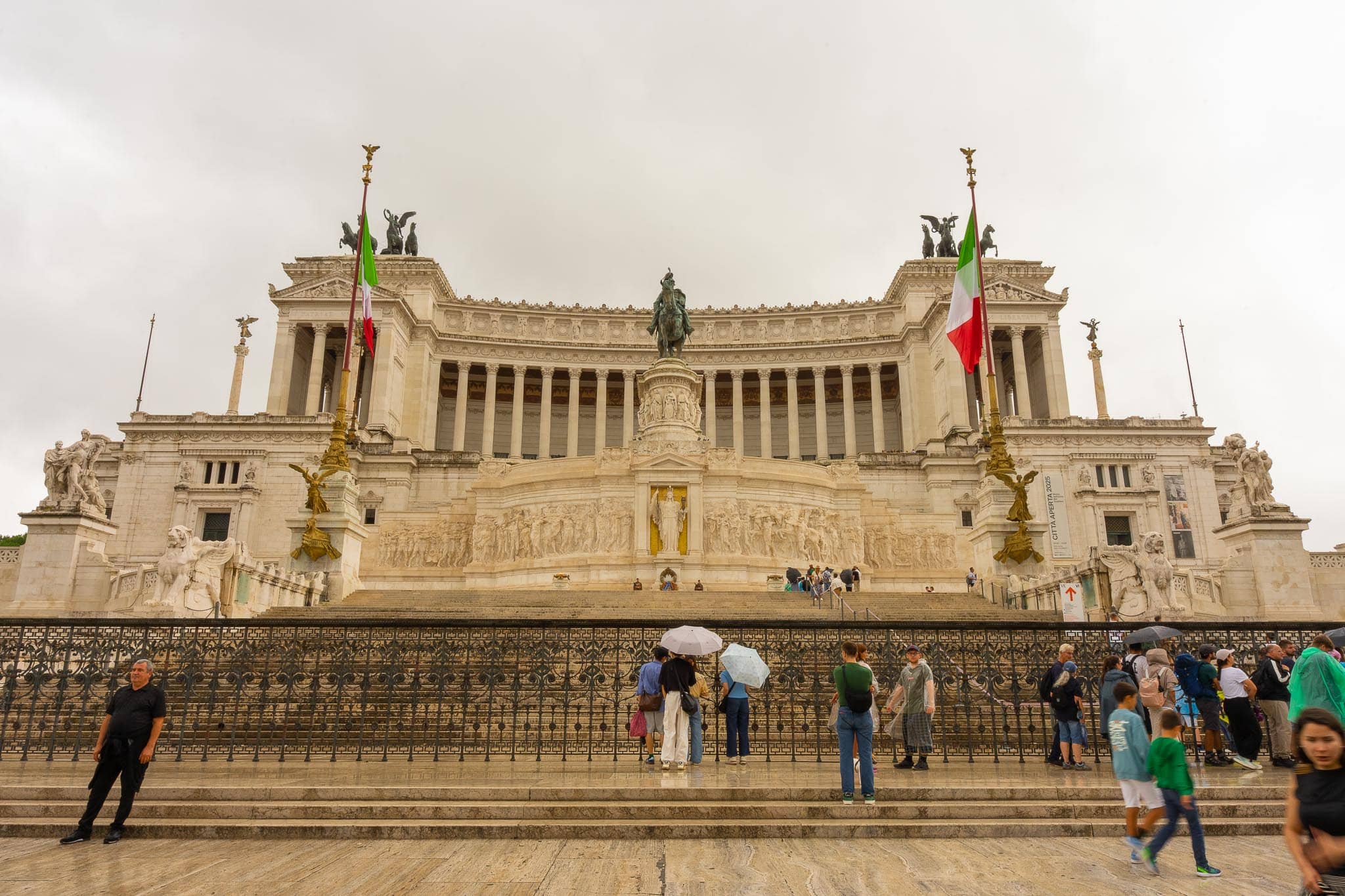 The white marble Vittoriano monument in Rome: colonnade, equestrian bronze and winged figures above a broad flight of steps, with Italian flags and visitors on the stairs.