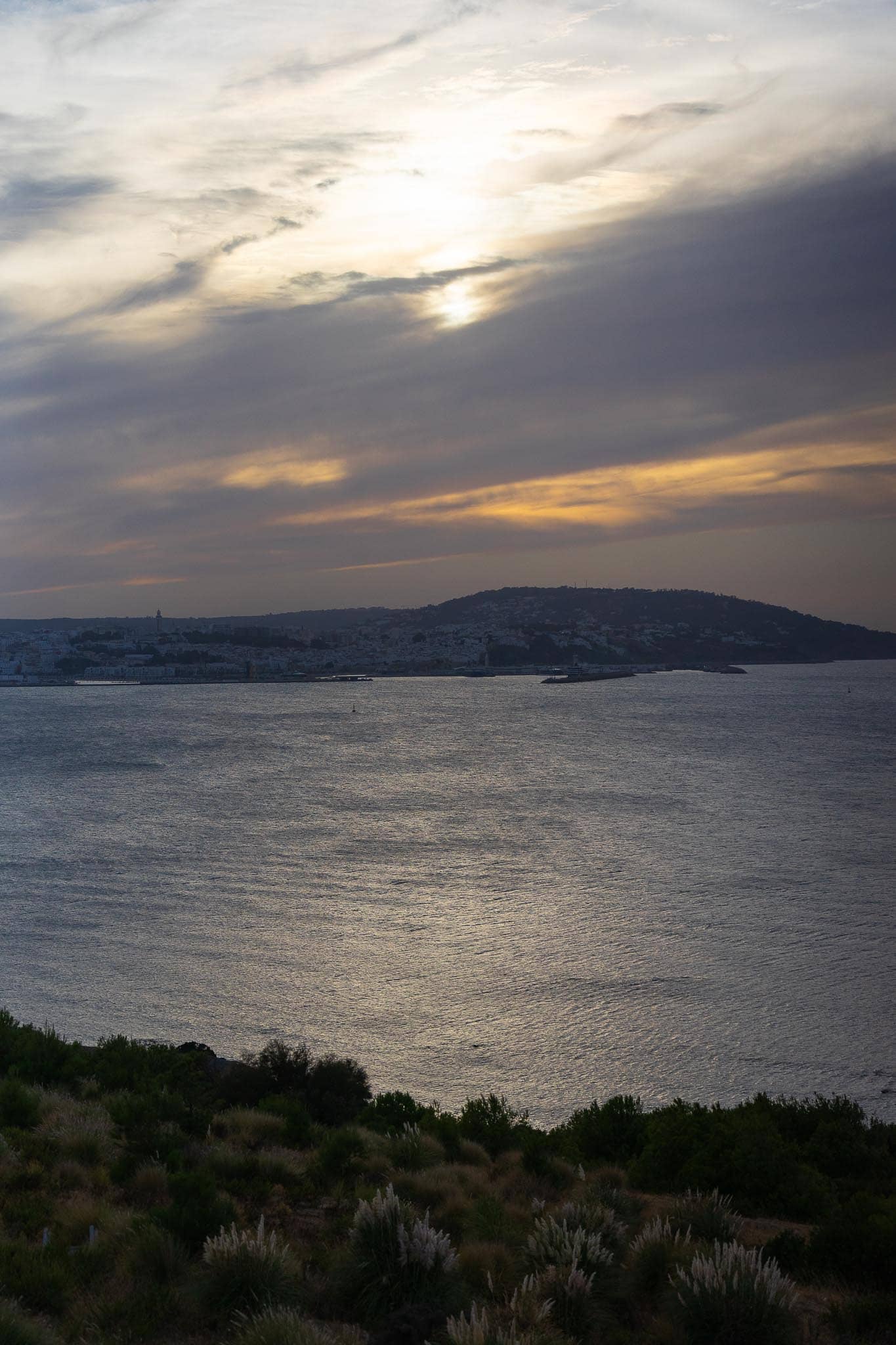 The Strait of Gibraltar at dusk from a scrubby hillside above Tangier: still grey water, a long headland, and the sun low behind heavy cloud.