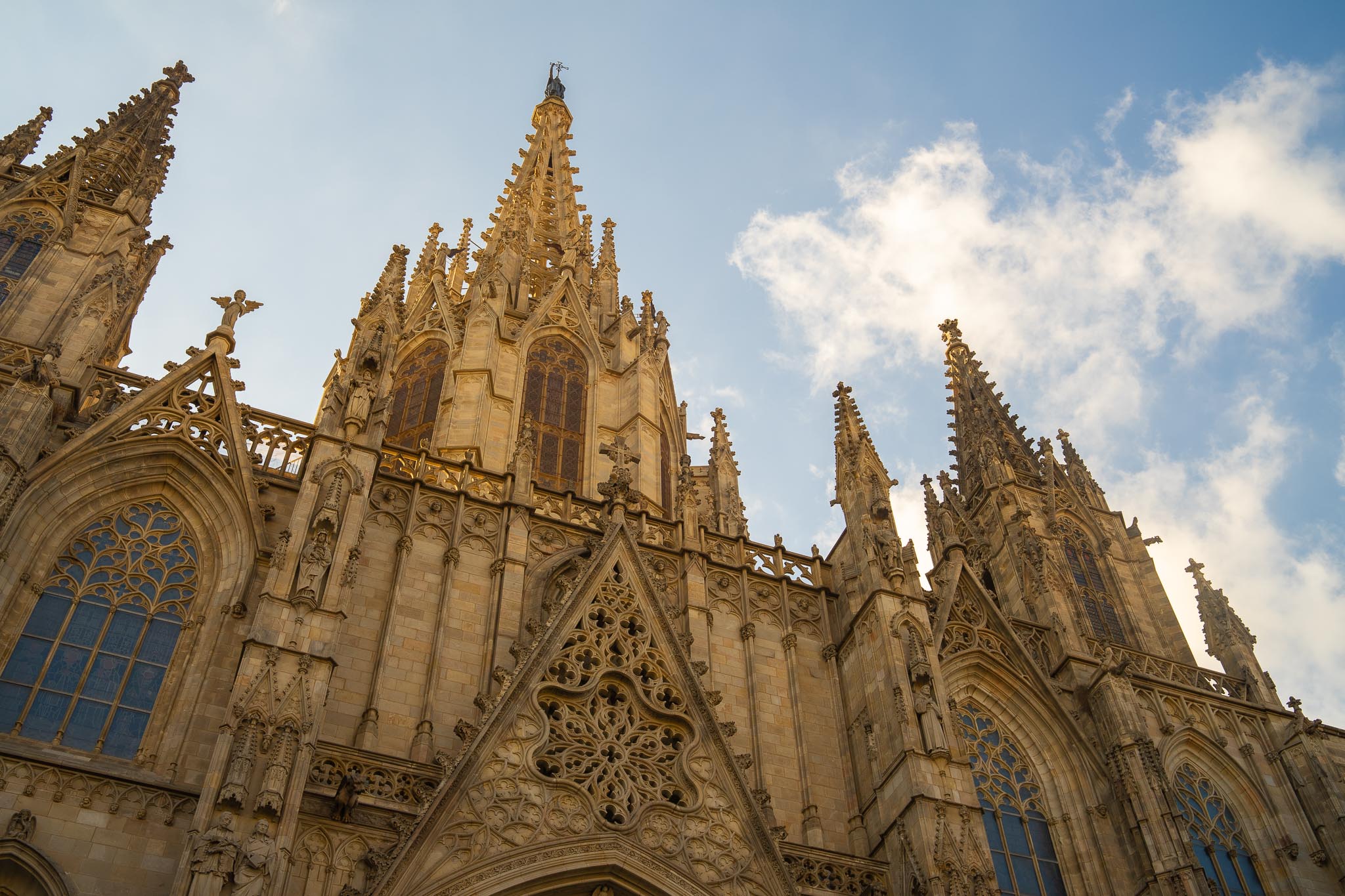 The upper front of Barcelona Cathedral seen steeply from below in warm low sunlight: a pale limestone gable of carved openwork tracery, a tall central spire and flanking pinnacles crowded with statues and crocketed finials, tall traceried windows on the left, and white cumulus in a blue sky behind.