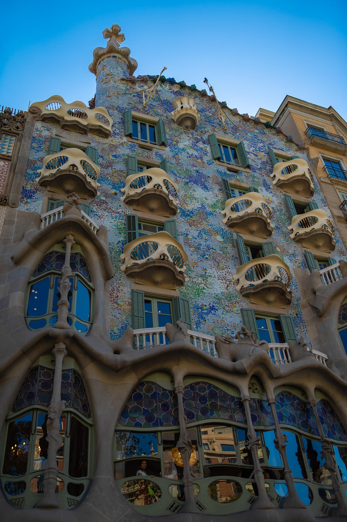 The façade of Casa Batlló on Passeig de Gràcia in Barcelona seen from the pavement below against a deep blue sky: a shimmering skin of blue, green and gold broken tile, bone-like stone columns and window frames on the lower storeys, rows of balconies shaped like masks, and a scaled roof ridge rising to a tiled turret topped with a cross.