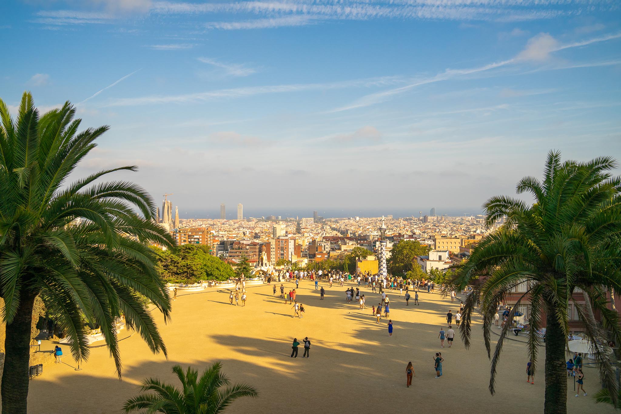 The wide sandy terrace of the Nature Square in Park Güell, Barcelona, framed by two tall date palms, with several dozen visitors scattered across it, a tiled pinnacle and the roofs of the entrance pavilions below, the city grid running away to the Sagrada Família and its cranes on the left, and the Mediterranean along the horizon under a pale blue sky.