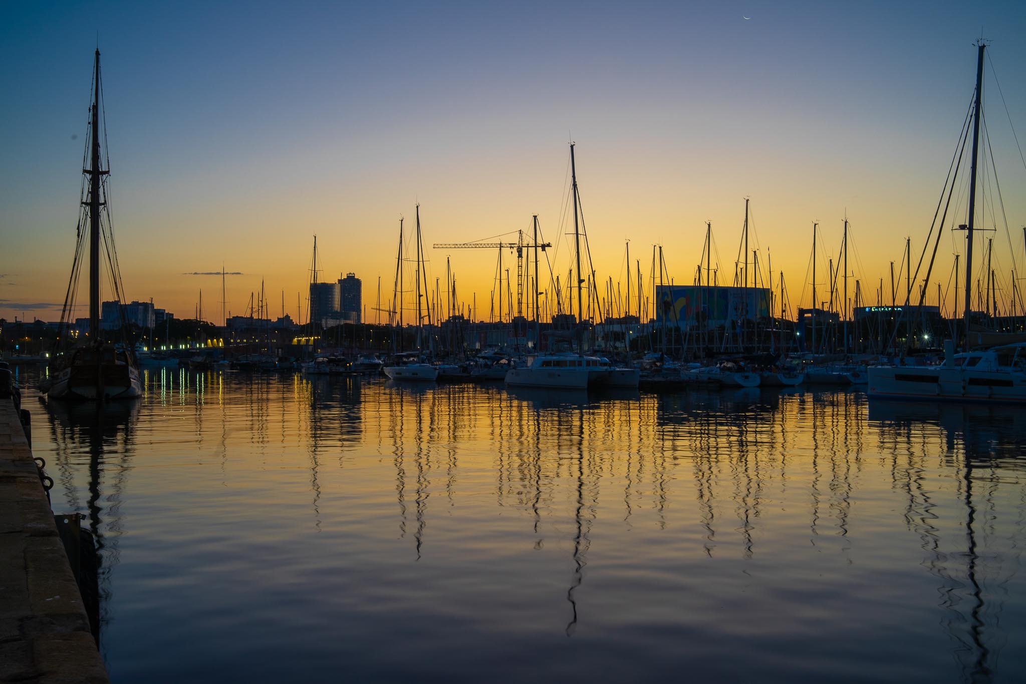 Port Vell in Barcelona under a low gold and deep blue sky: dozens of yacht masts silhouetted along the far quay with their reflections running down the still black water, a thin crescent moon high on the right, a construction crane and the towers of the seafront on the horizon, and the stone edge of the wharf in the foreground.
