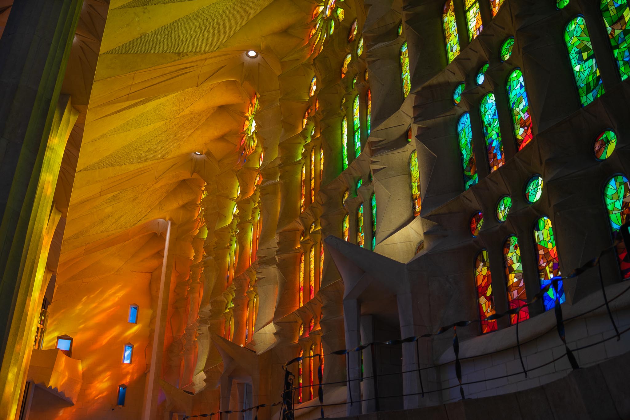 Inside the Sagrada Família in Barcelona: pale stone piers and a folded, faceted wall washed in deep orange and yellow light from unseen windows on the left, and on the right a tall wall of stained glass in greens, blues and reds above a curved stone balcony.