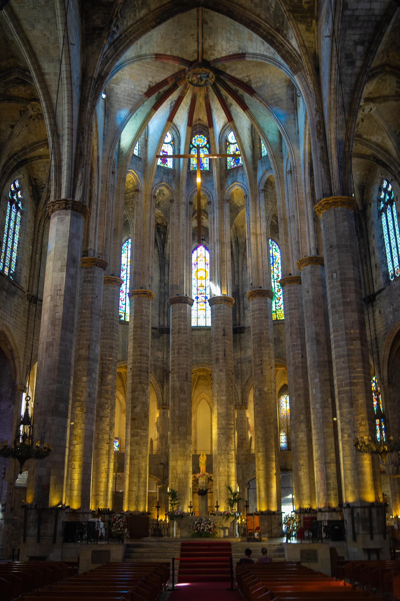 The interior of Santa Maria del Mar in Barcelona looking east to the apse: tall plain octagonal stone columns set far apart, rising to a ribbed vault washed with pale light, a suspended cross on a long chain above the crossing, stained glass in the clerestory and ambulatory, floodlit stone and rows of dark wooden pews below.