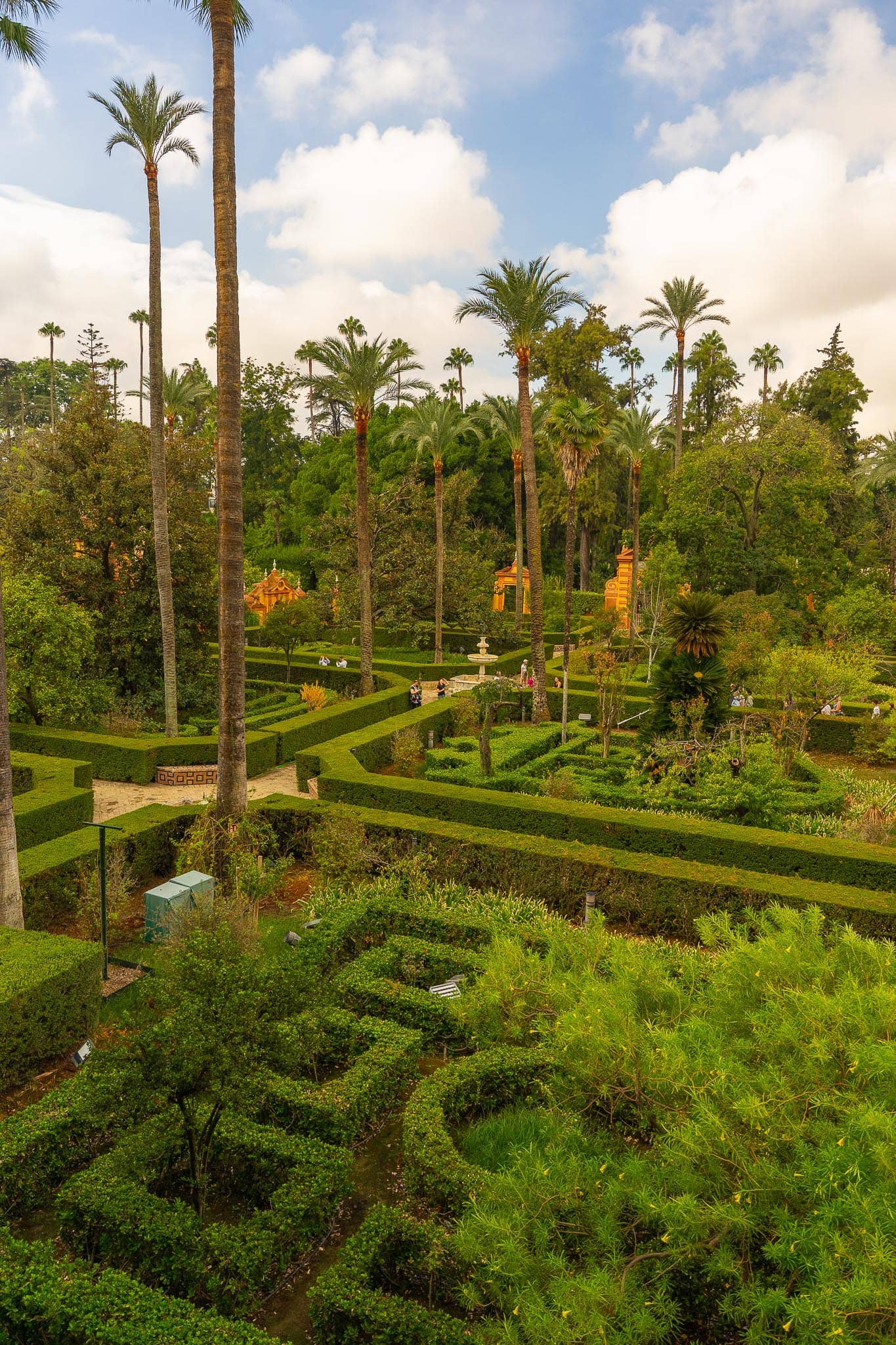 The gardens of the Real Alcázar in Seville seen from above: a grid of clipped box parterres and gravel paths, very tall Canary palms, an ochre garden pavilion among dark trees, a small stone fountain, and a few visitors on the paths.