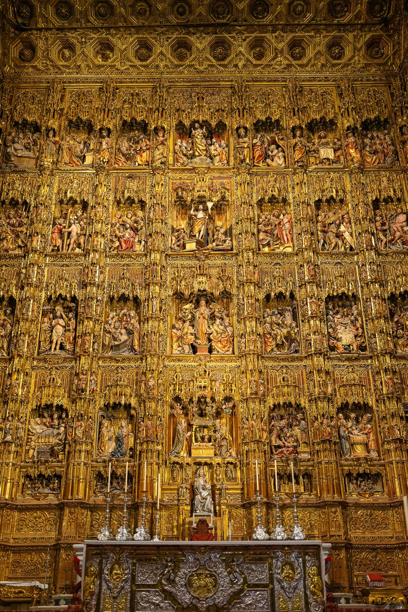 The Retablo Mayor of Seville Cathedral: a vast gilded altarpiece rising in tiers of carved and painted scenes beneath a coffered gold canopy, with silver candlesticks and an ornate silver altar frontal below.