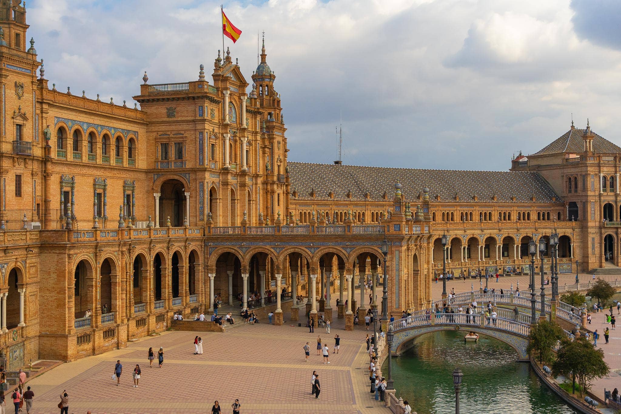 Plaza de España in Seville seen from an elevated position on its arcade: the tiled and pinnacled north tower flying a Spanish flag, the long brick and azulejo colonnade, the canal with a rowing boat passing under a balustraded tiled bridge, and several dozen visitors on the paving below.